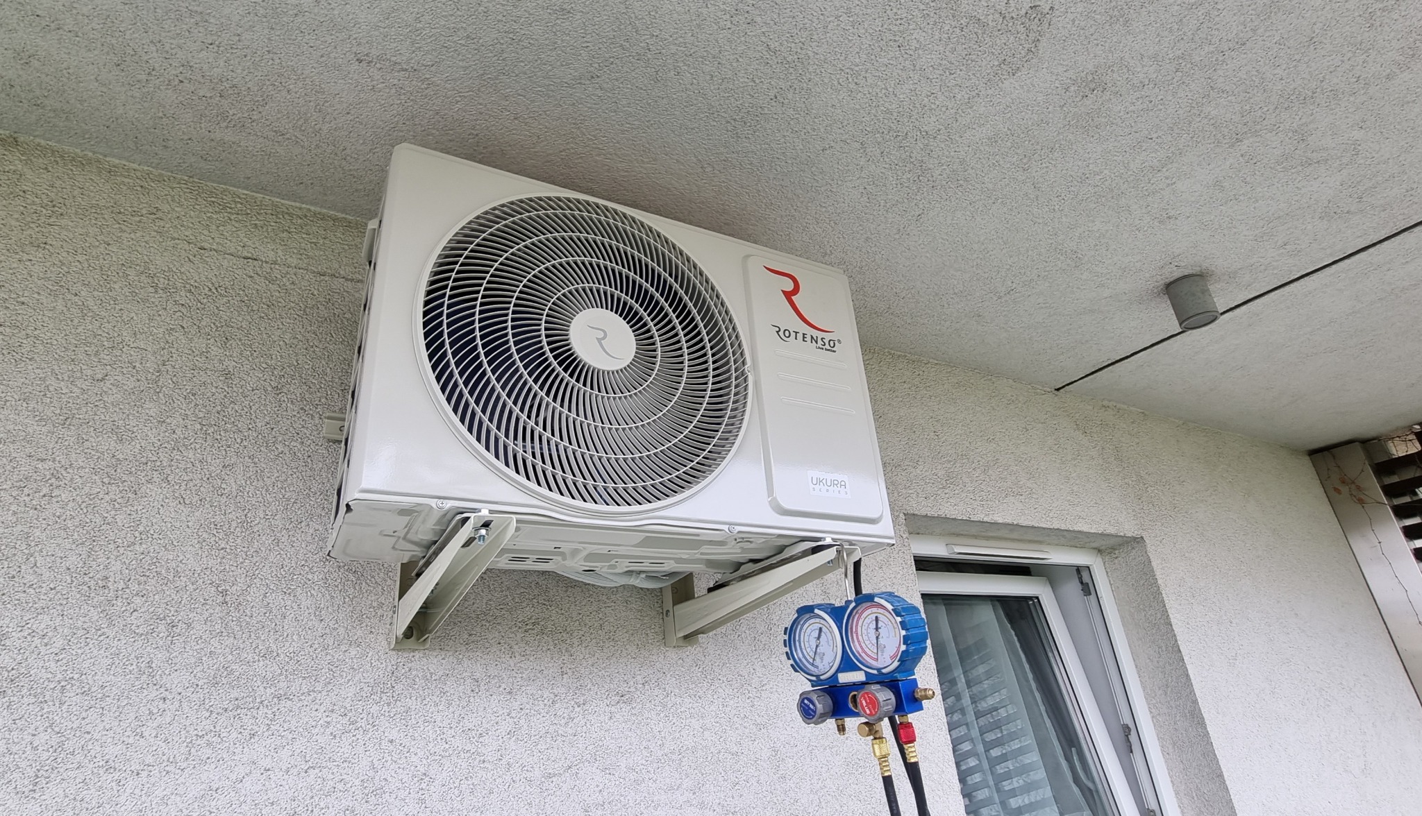 Biała jednostka klimatyzacji Rotenso Ukura zamontowana na zewnątrz budynku, podłączona do manometru serwisowego z wężykami, widok z dołu.