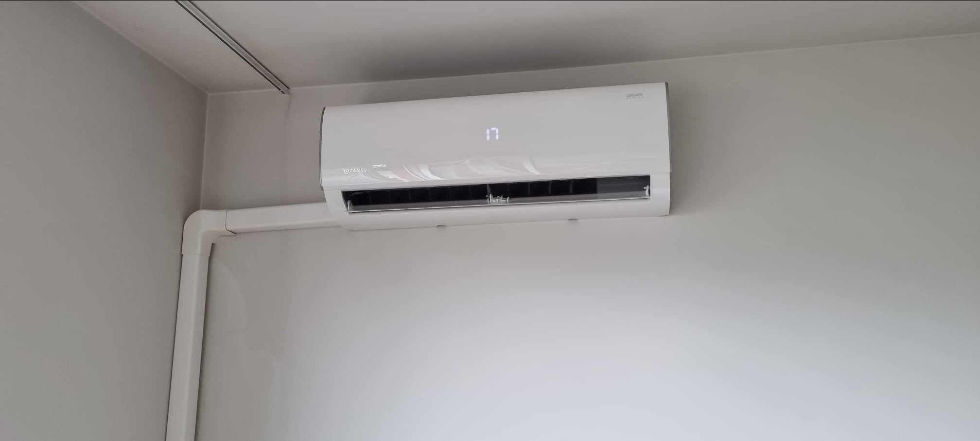 Biały klimatyzator Rotenso zamontowany na jasnoszarej ścianie, z widocznymi białymi rurami instalacyjnymi i wyświetlaną temperaturą 17 stopni.
