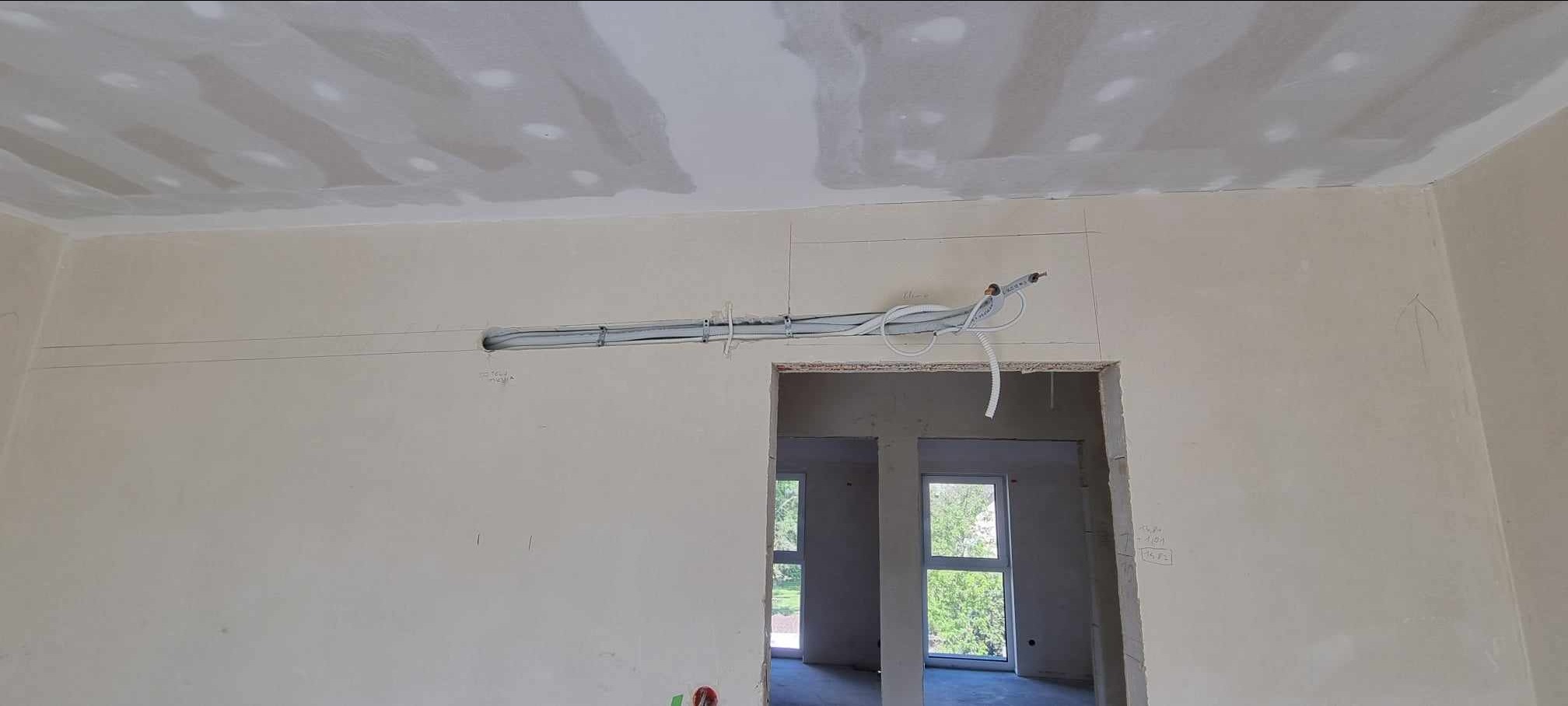 Wnętrze w trakcie montażu instalacji klimatyzacyjnej; widoczne rury prowadzone pod sufitem, ściany w stanie surowym, przygotowane do dalszych prac wykończeniowych, otwór w ścianie na przewody.