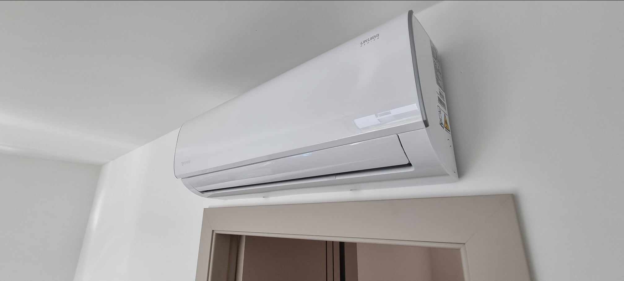 Biała jednostka wewnętrzna klimatyzacji marki Ukura zamontowana na ścianie nad futryną drzwiową w jasnym pomieszczeniu.