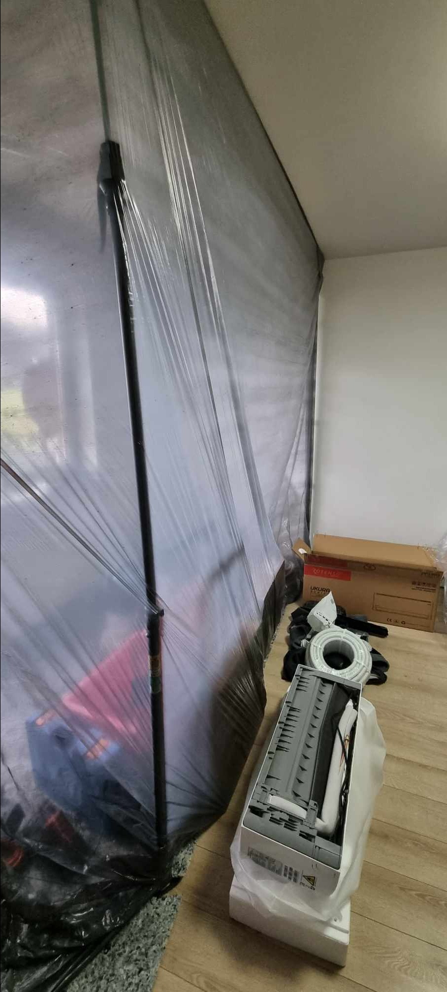 Przygotowania do montażu klimatyzacji w pomieszczeniu, ściany zabezpieczone folią, rozpakowany klimatyzator w kartonie, zwój kabla.