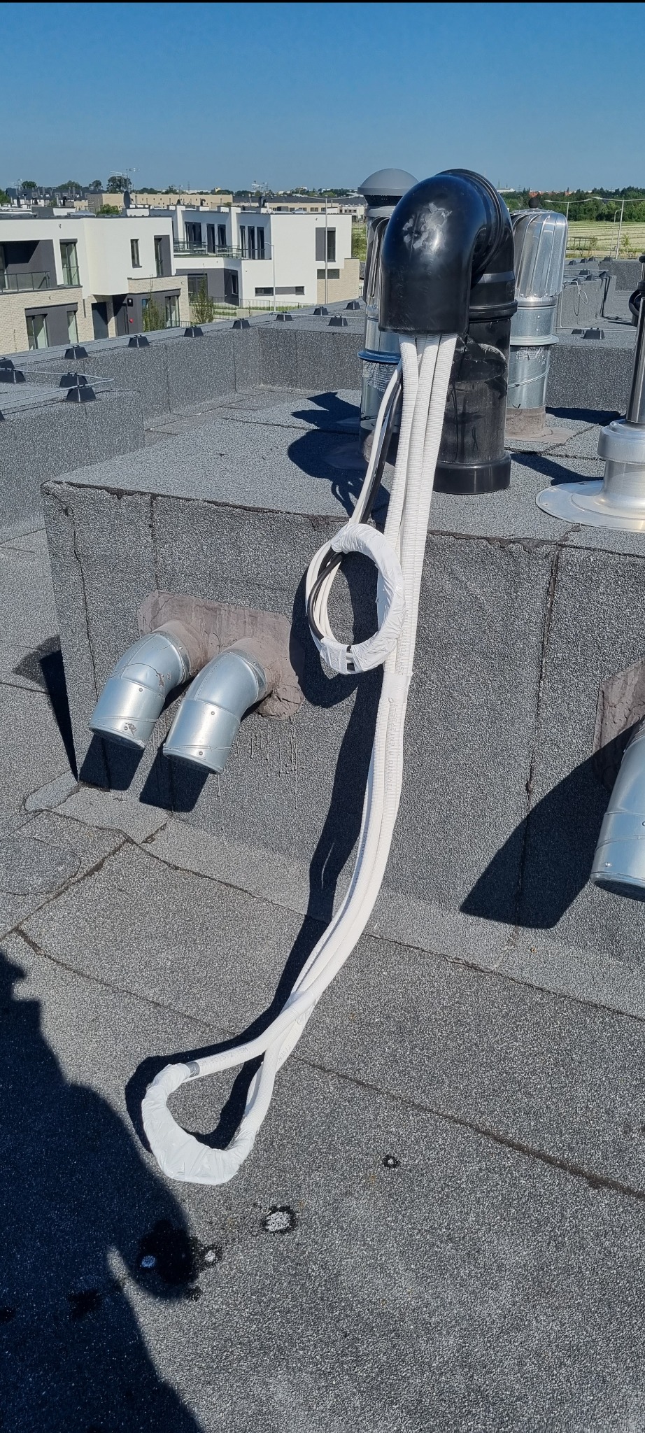 Instalacja klimatyzacji na dachu budynku mieszkalnego, widoczne rury odprowadzające i przewody w białej izolacji, na tle zabudowa osiedlowa w słoneczny dzień.