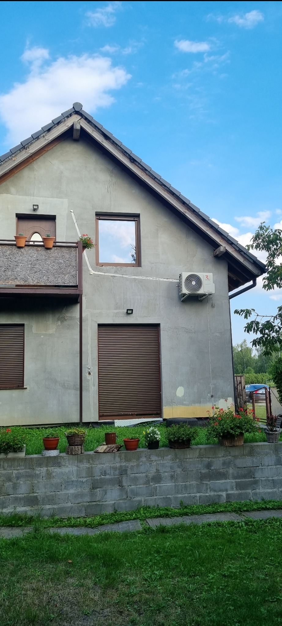 Szary dom jednorodzinny z zamontowaną klimatyzacją na ścianie, widoczne brązowe rolety, balkon z kwiatami w doniczkach i fragment ogrodu z zielenią.