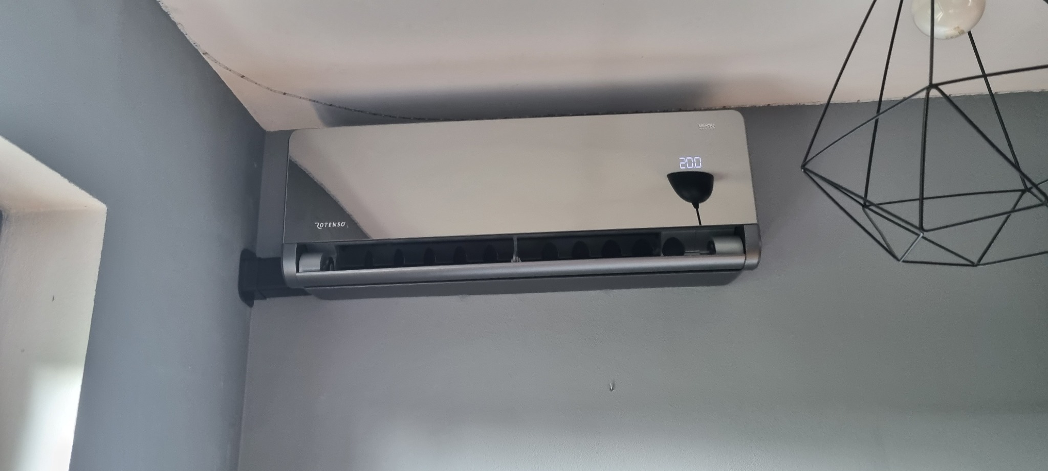 Szary klimatyzator Rotenso zamontowany na szarej ścianie w pomieszczeniu, widoczny wyświetlacz wskazujący temperaturę 20 stopni Celsjusza, obok nowoczesna lampa sufitowa.