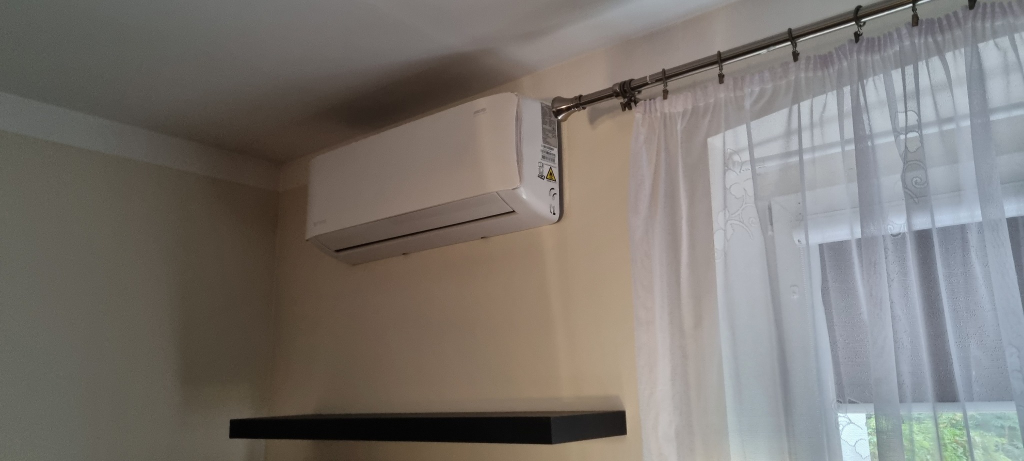 Biały klimatyzator ścienny zamontowany w pokoju z kremowymi ścianami, widoczna czarna półka i okno z białą, wzorzystą firanką zawieszoną na metalowym karniszu.