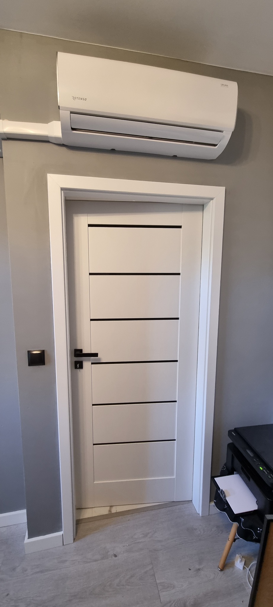 Zamontowana klimatyzacja ROTENSO na szarej ścianie nad białymi drzwiami z czarnymi paskami, widoczne elementy instalacji elektrycznej i fragment drukarki.