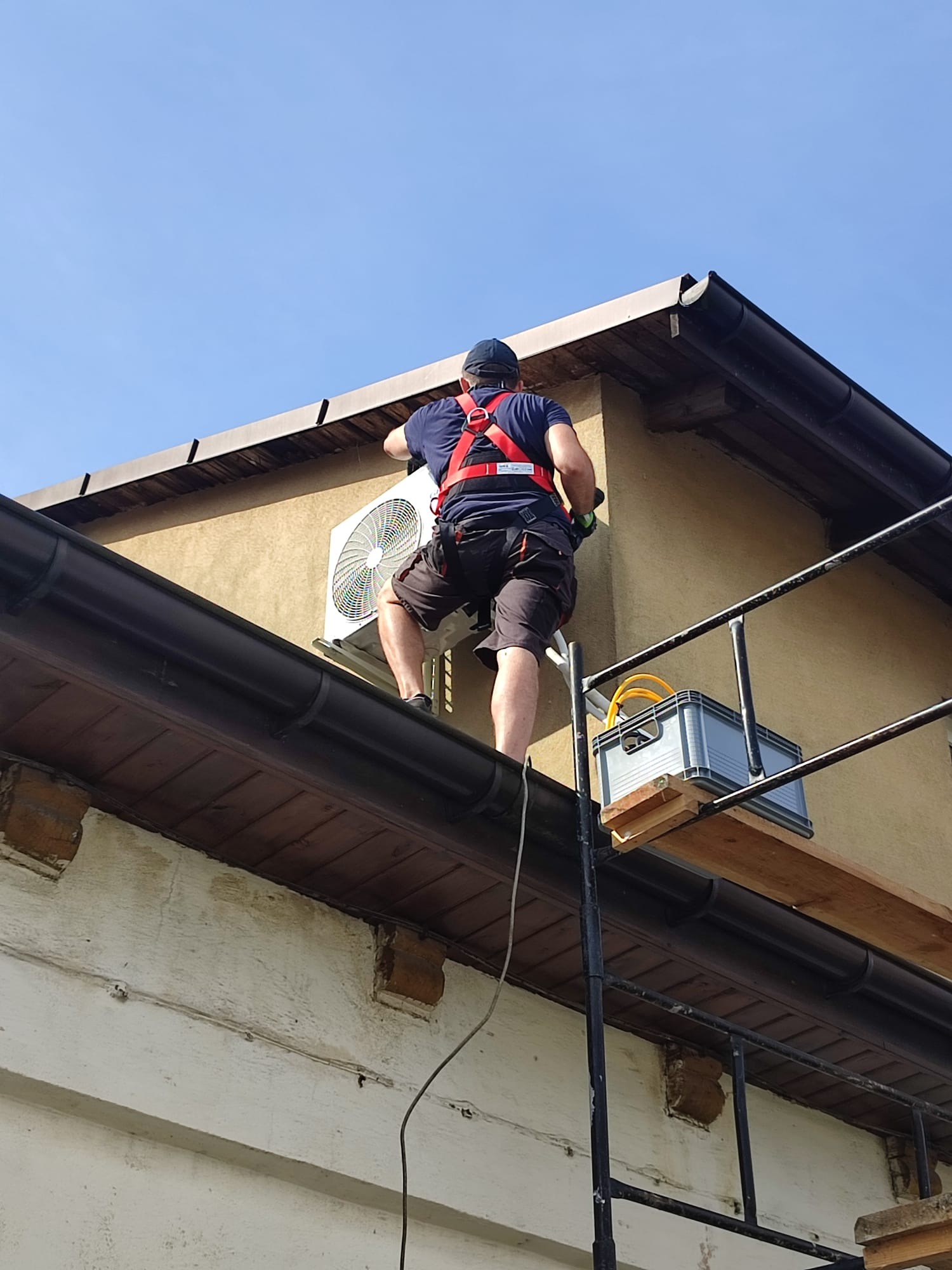 Mężczyzna w szelkach asekuracyjnych montuje zewnętrzną jednostkę klimatyzatora na ścianie budynku, stojąc na rusztowaniu z szarym pojemnikiem na narzędzia.