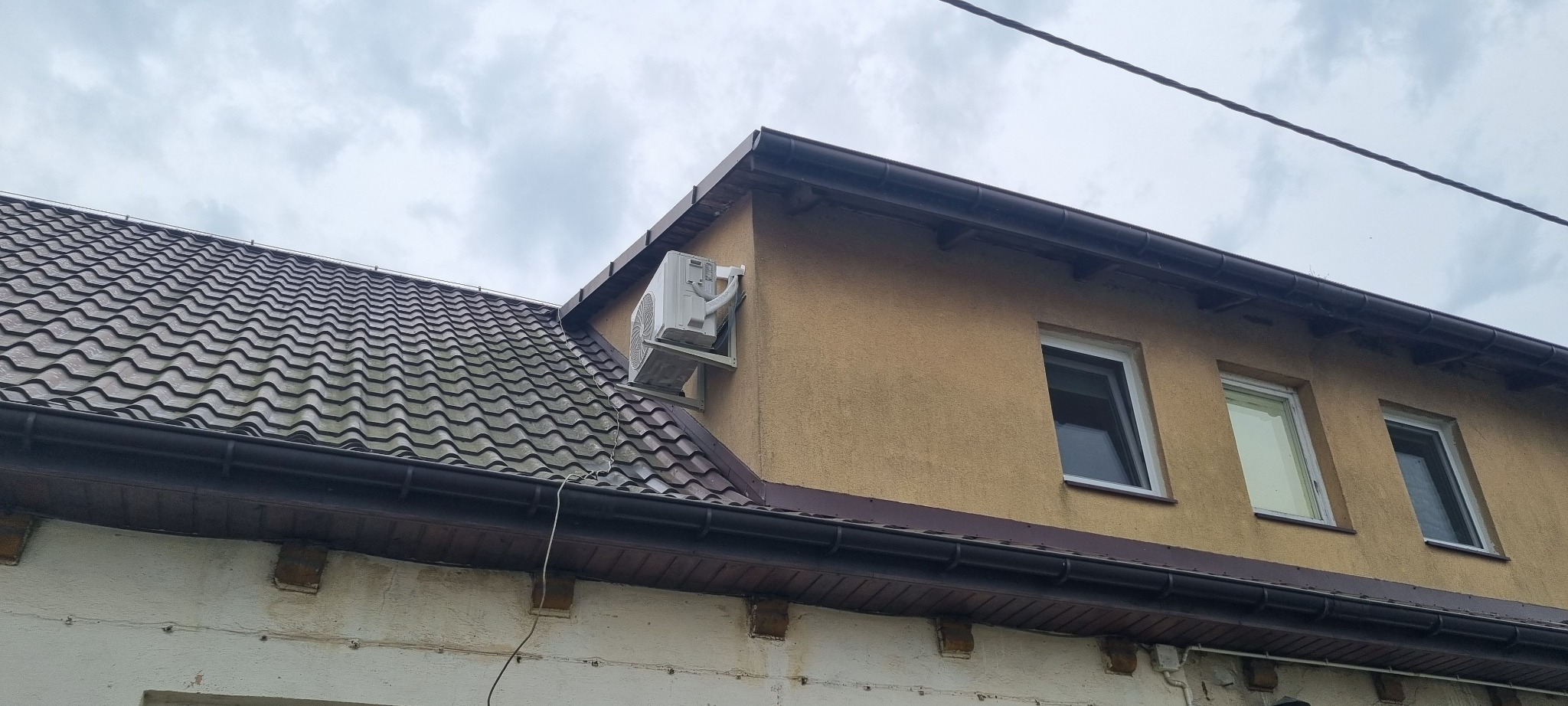 Zewnętrzna jednostka klimatyzacji zamontowana na ścianie domu z brązową dachówką, widok z dołu.