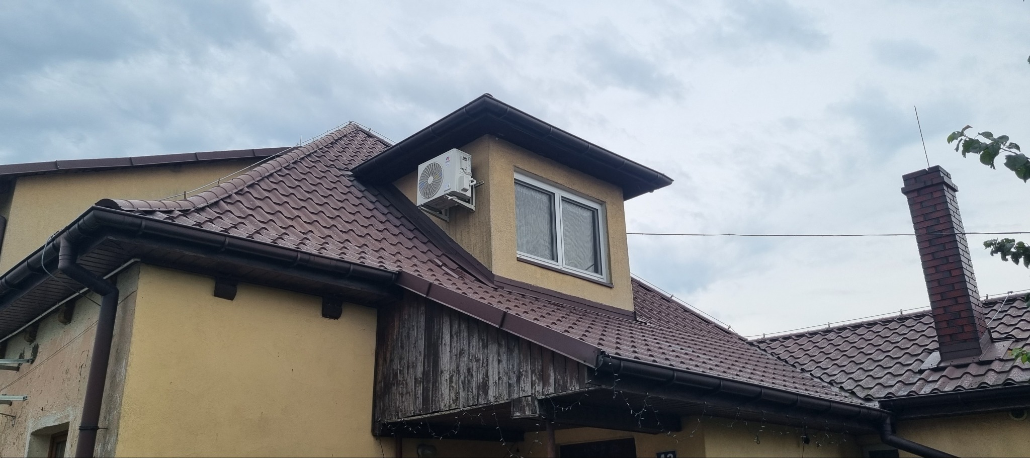 Zewnętrzna jednostka klimatyzacji zamontowana na ścianie domu z brązowym dachem, widoczny komin i okno poddaszowe, pochmurne niebo w tle.