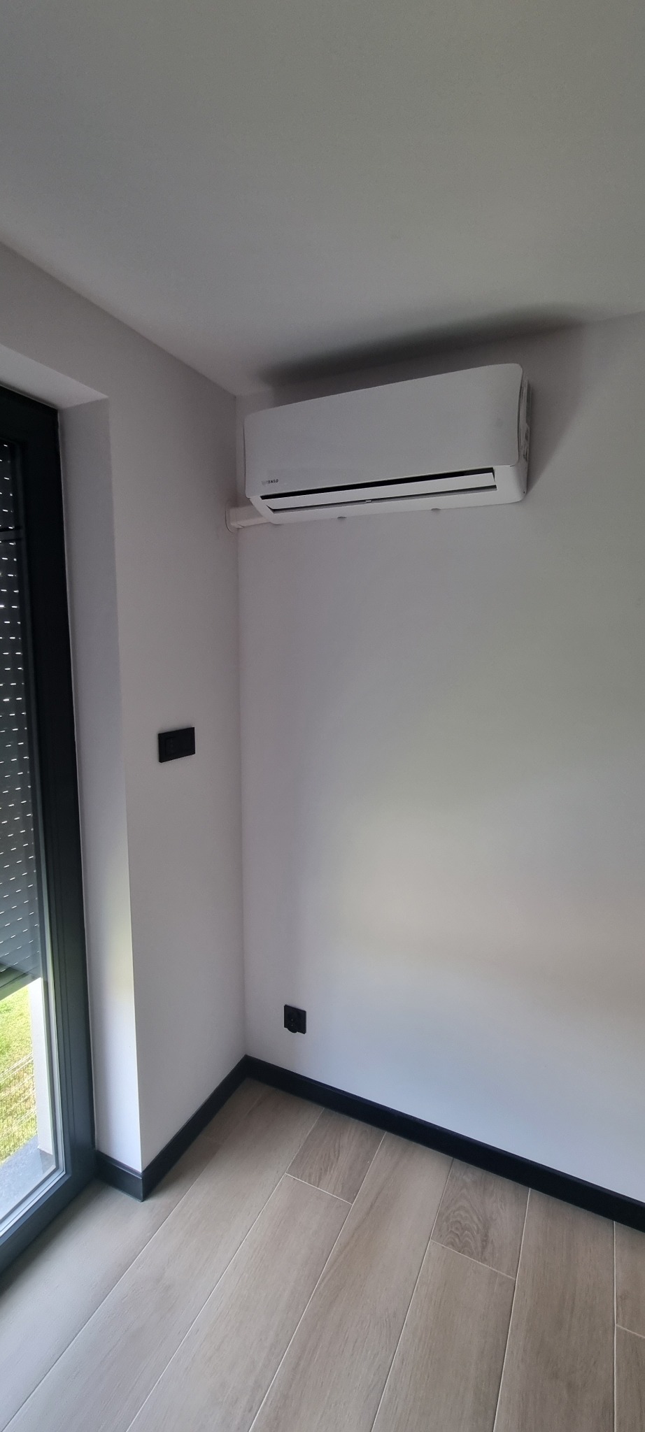 Nowoczesny, biały klimatyzator ścienny marki Tenso, zamontowany w rogu jasnego pokoju z drewnopodobną podłogą i czarnymi listwami przypodłogowymi.