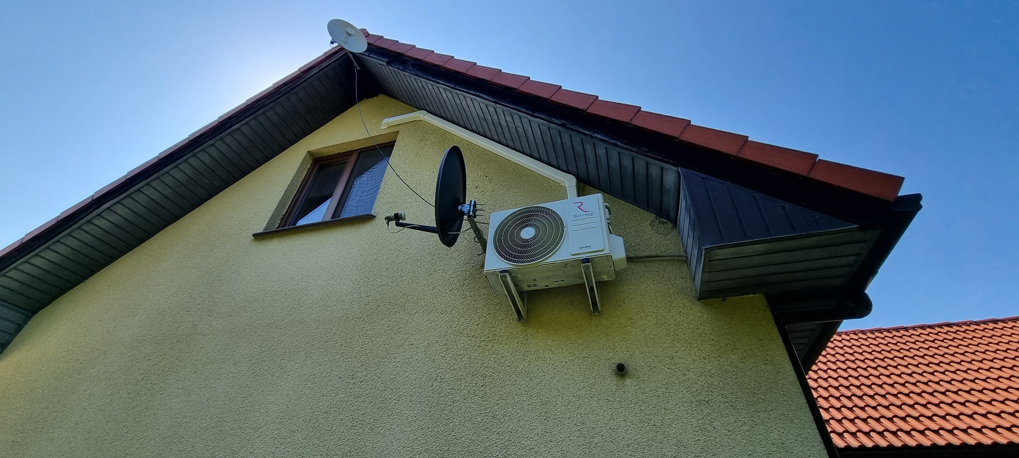 Zewnętrzna jednostka klimatyzacji Rotenso zamontowana na elewacji domu, obok okna i anteny satelitarnej, widok z dołu.