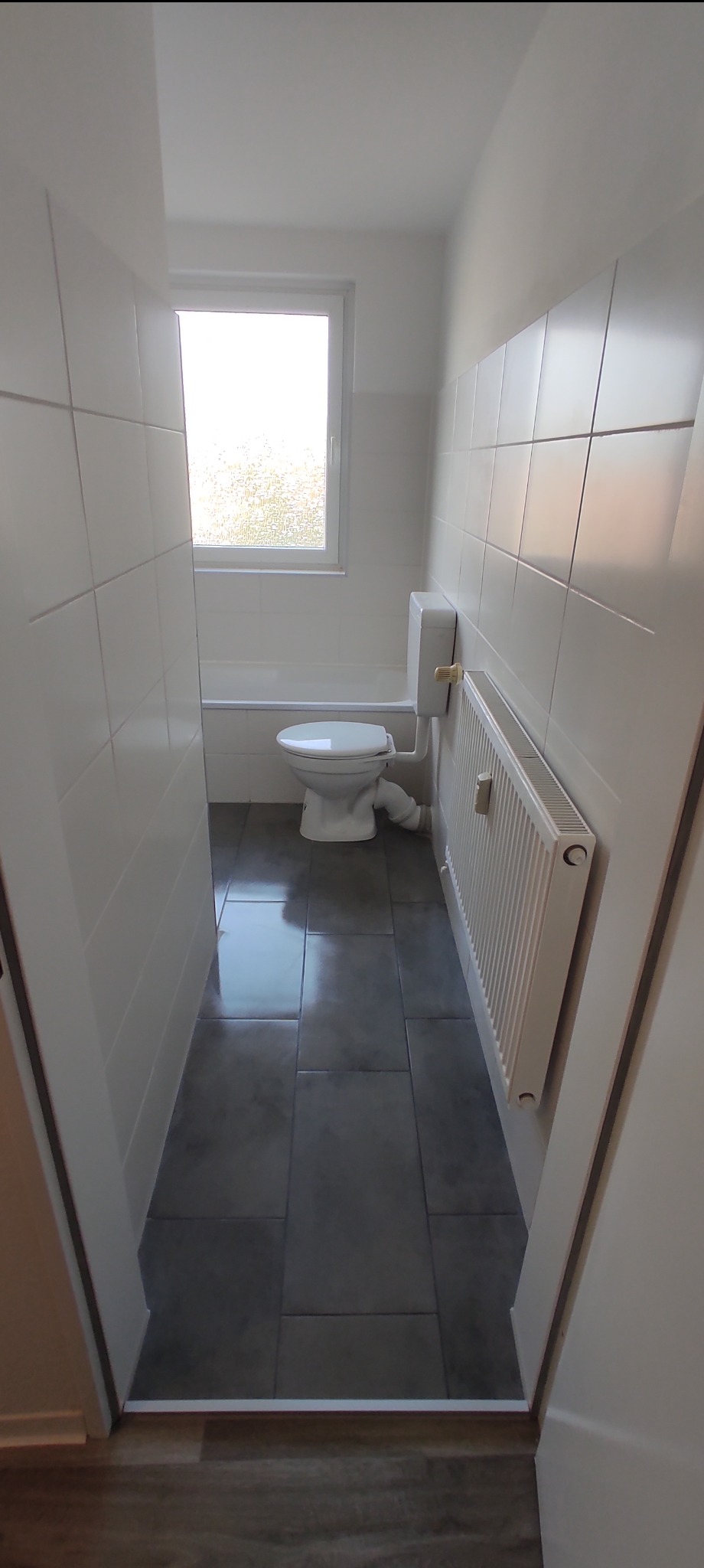 Wąskie ujęcie wyremontowanej łazienki z białymi płytkami, szarą podłogą, grzejnikiem i toaletą pod oknem, widok zza framugi drzwi.
