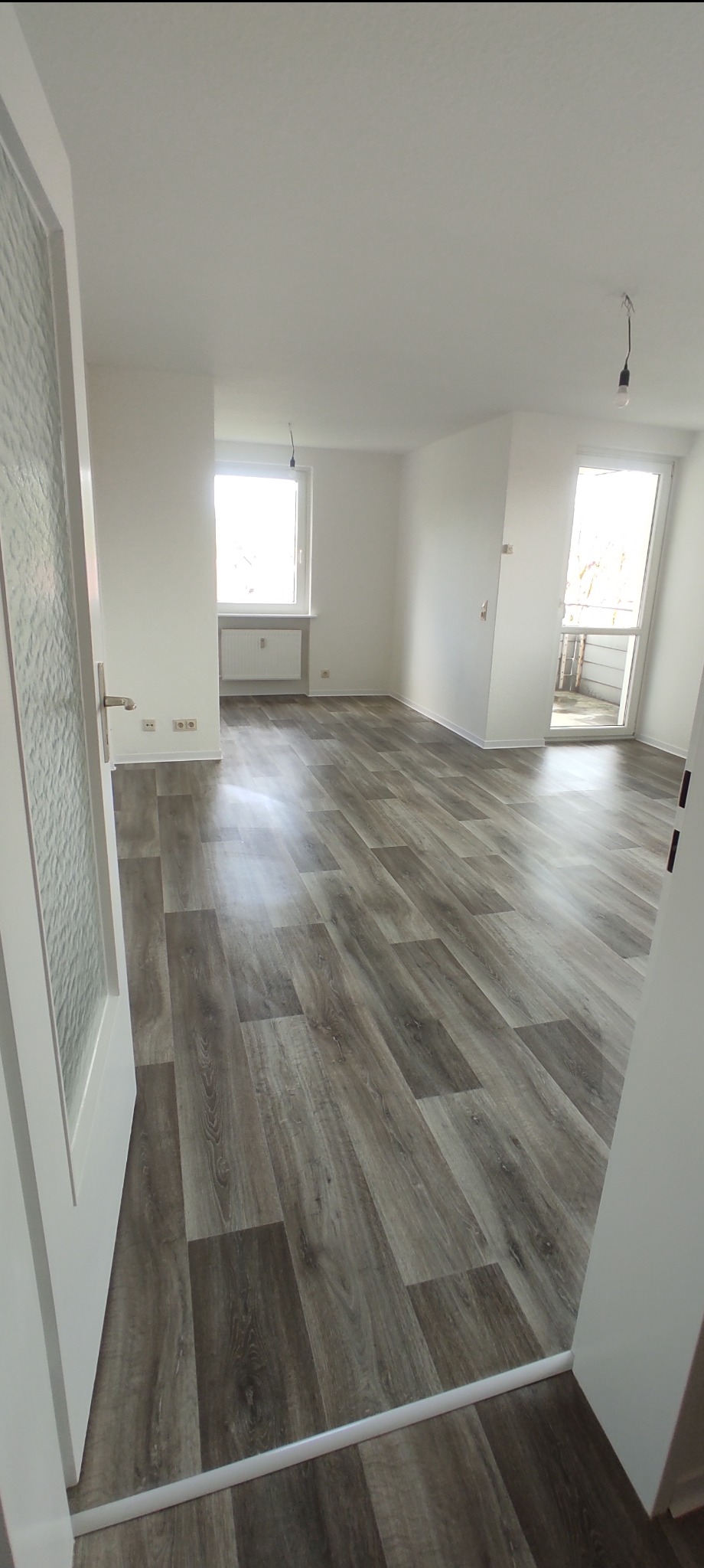 Pusty pokój po remoncie z panelami podłogowymi w odcieniach szarości, białe ściany, okno i drzwi balkonowe, widok zza uchylonych drzwi z mleczną szybą.