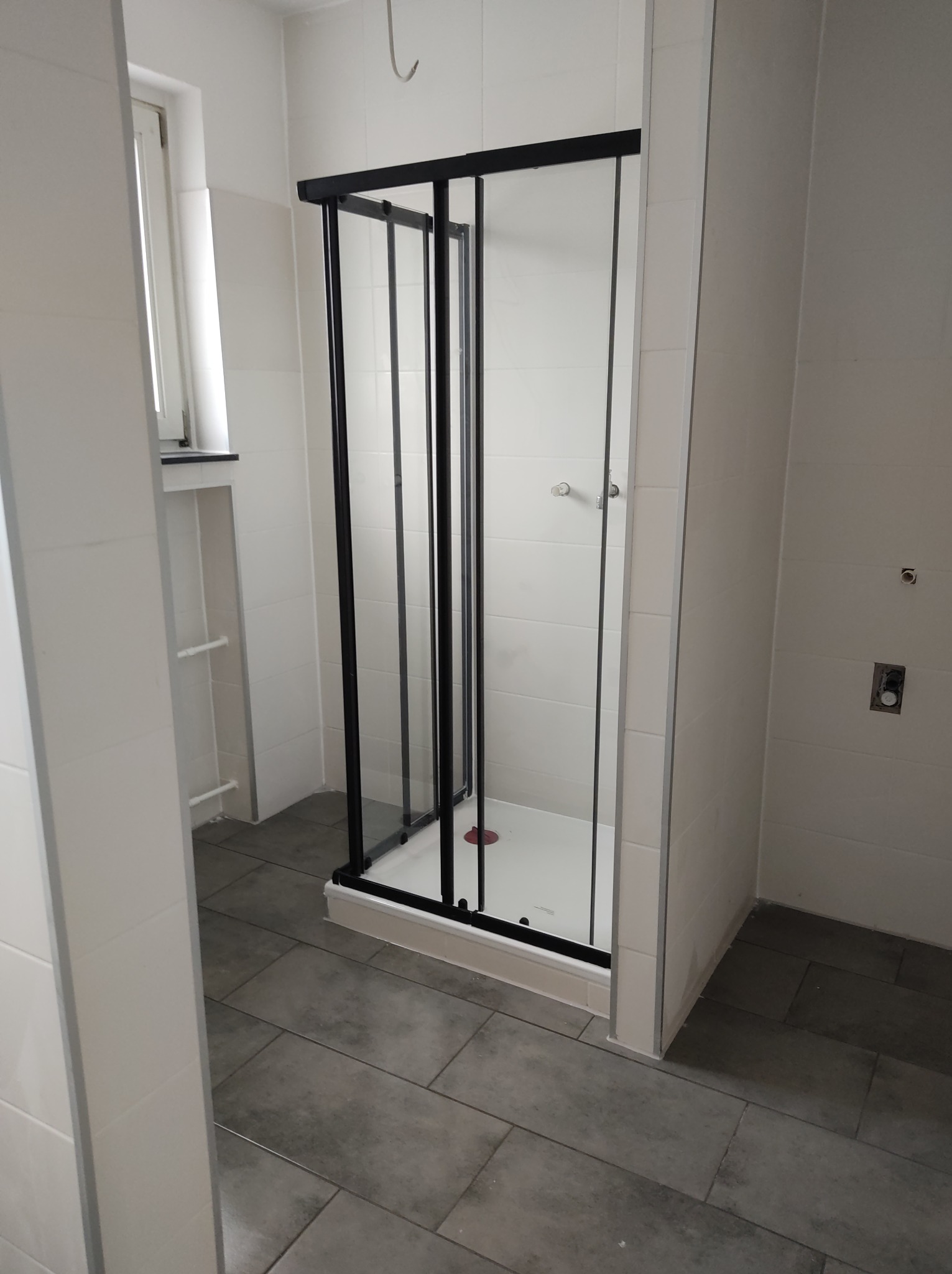 Nowoczesna kabina prysznicowa z czarnymi profilami i przesuwnymi drzwiami w jasnej łazience z szarą podłogą i białymi płytkami ściennymi.