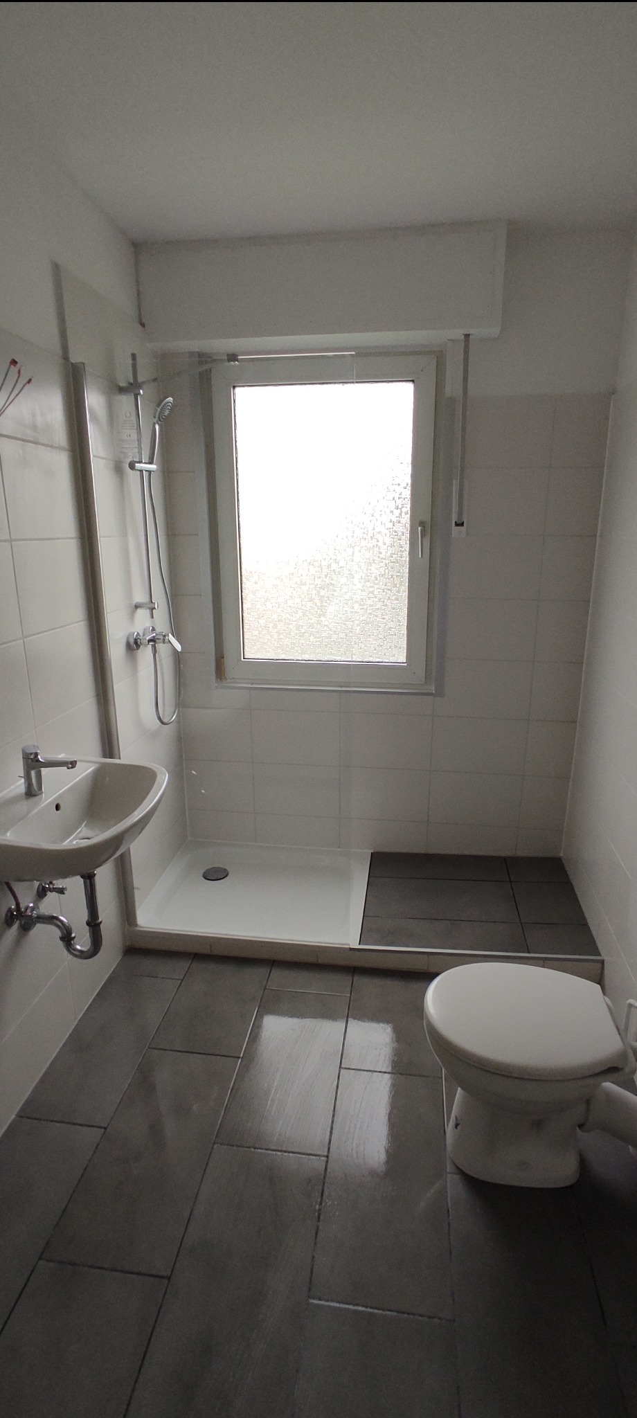 Wykończona łazienka z szarymi płytkami podłogowymi, białą kabiną prysznicową, umywalką i toaletą, oświetlona naturalnym światłem z okna z szybą ornamentową.