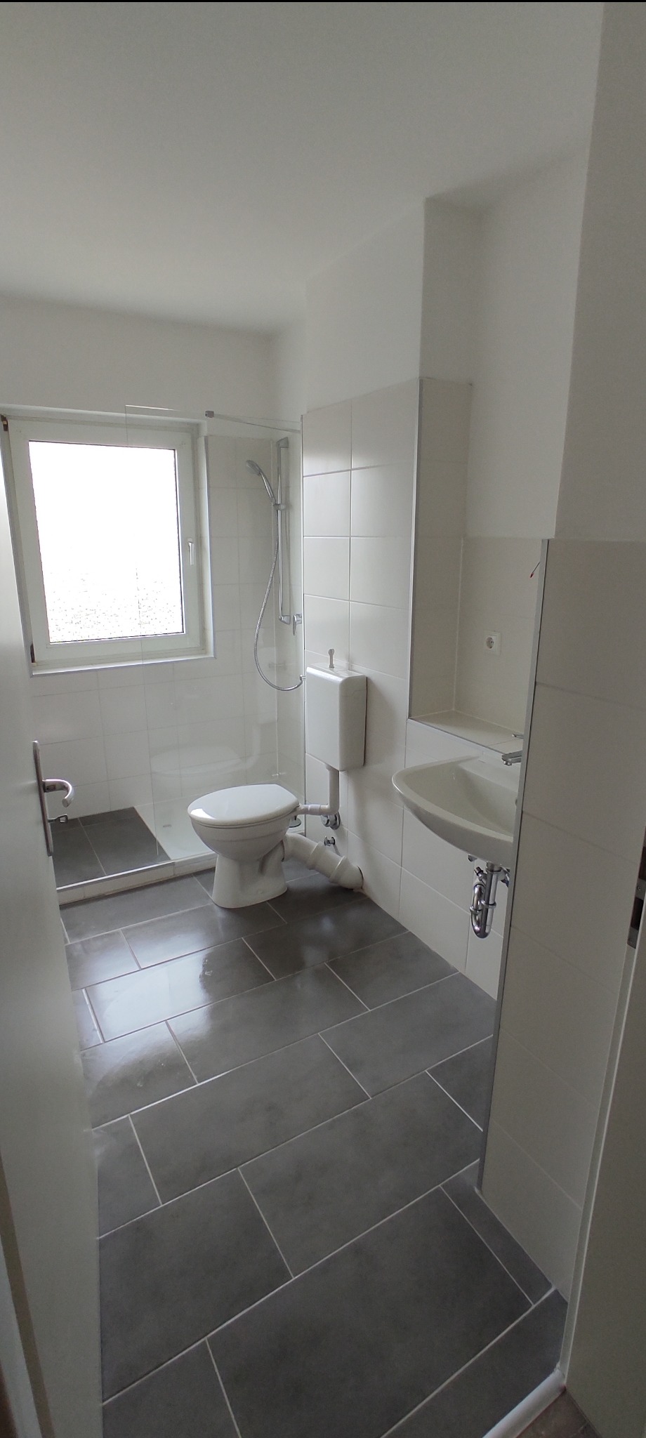 Nowoczesna łazienka z szarymi płytkami podłogowymi ułożonymi w prostokąty, białą armaturą i kabiną prysznicową z przezroczystego szkła.