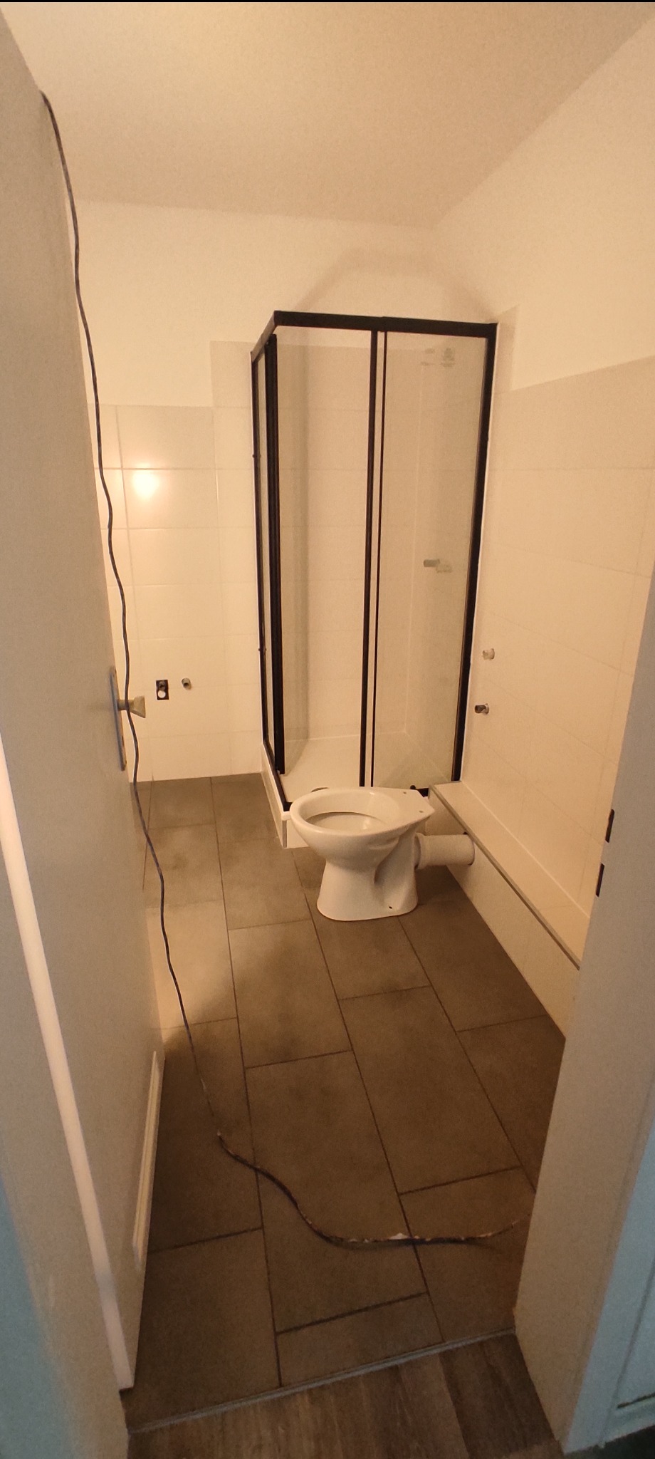 Niedokończona łazienka z kabiną prysznicową z czarnymi ramami i toaletą bez spłuczki, brązowe płytki podłogowe, widoczne kable na podłodze i fragment drzwi.