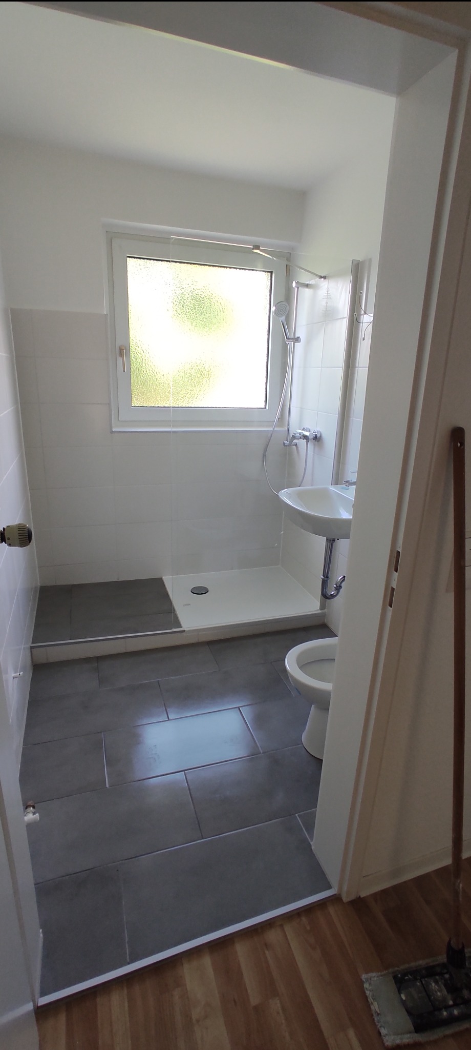 Nowoczesna łazienka z szarą podłogą, białymi płytkami, kabiną prysznicową bez brodzika i wiszącą umywalką, widok zza futryny drzwi, mop na podłodze.