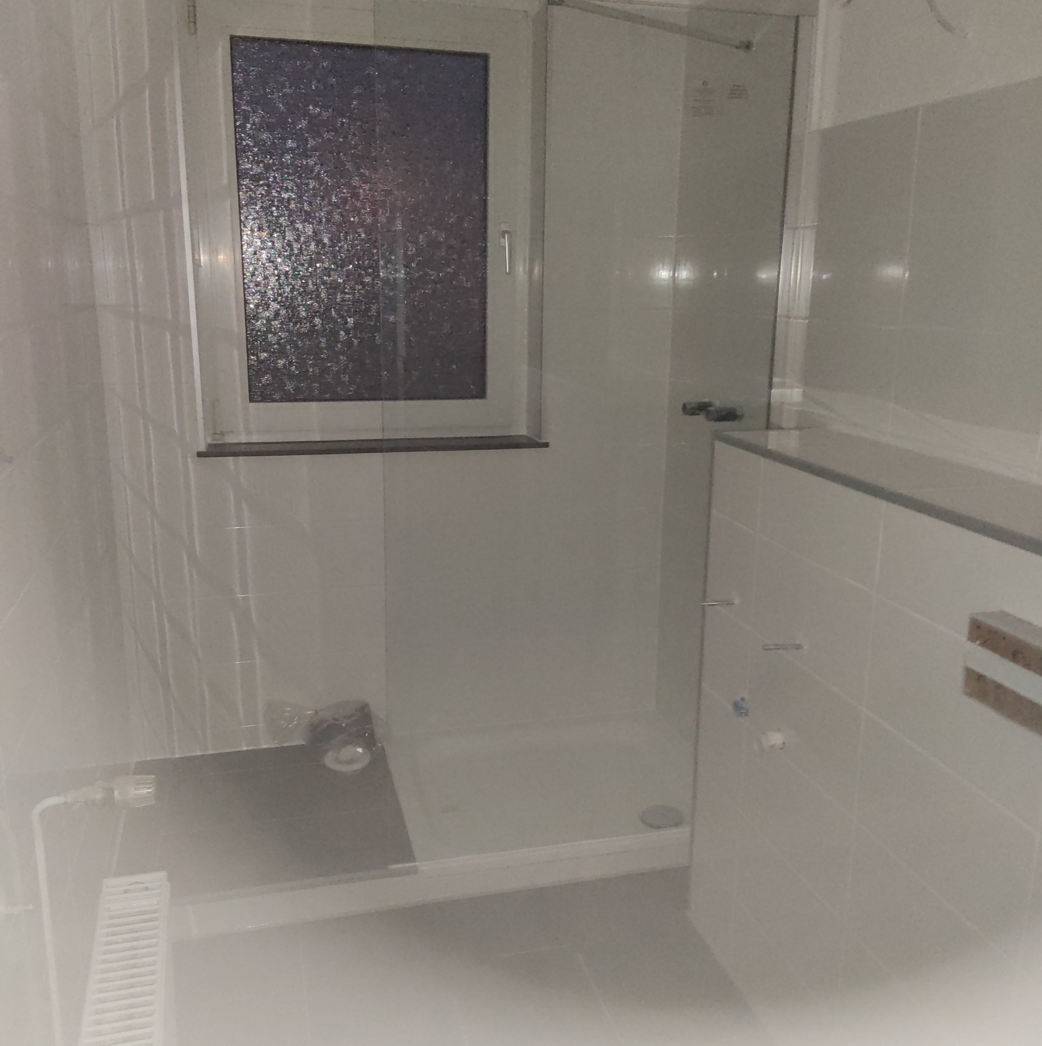 Nowoczesna łazienka z kabiną prysznicową, białe płytki, okno z ozdobną szybą, grzejnik żeberkowy i elementy armatury.