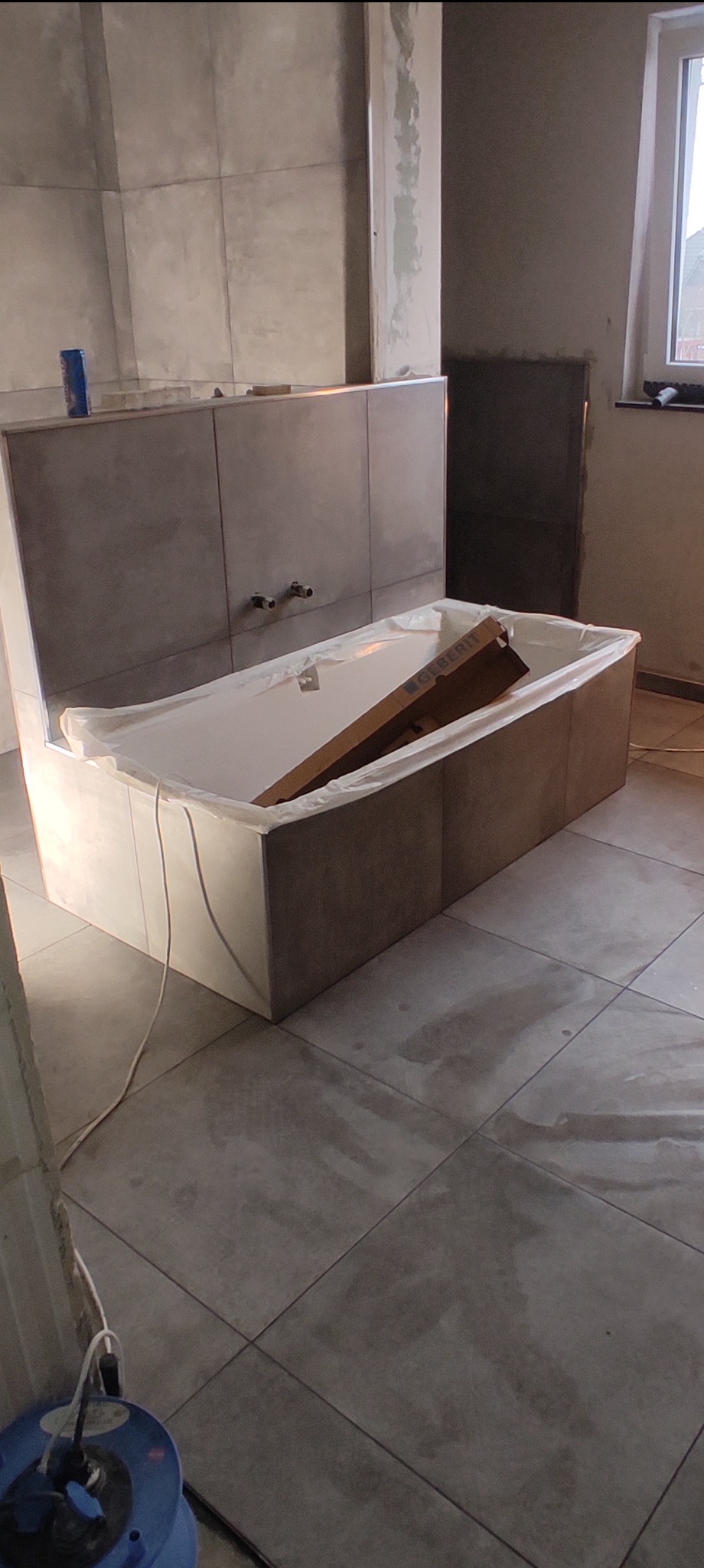 Wanna obudowana płytkami w trakcie montażu, zabezpieczona folią i kartonem Geberit, widoczne przygotowane przyłącza wodne na ścianie wyłożonej płytkami, na podłodze płytki w odcieniach szarości...