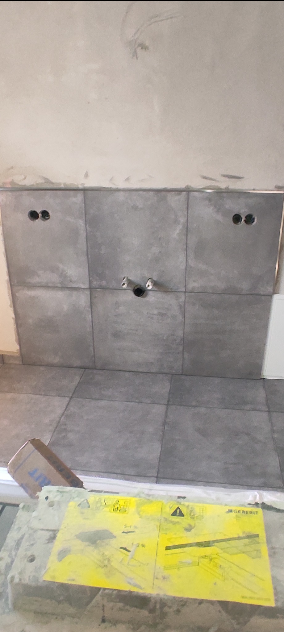 Szara, matowa glazura ścienna i podłogowa w łazience, z widocznymi otworami na instalacje sanitarne i elektryczne, oraz żółta instrukcja montażu Geberit na pierwszym planie.