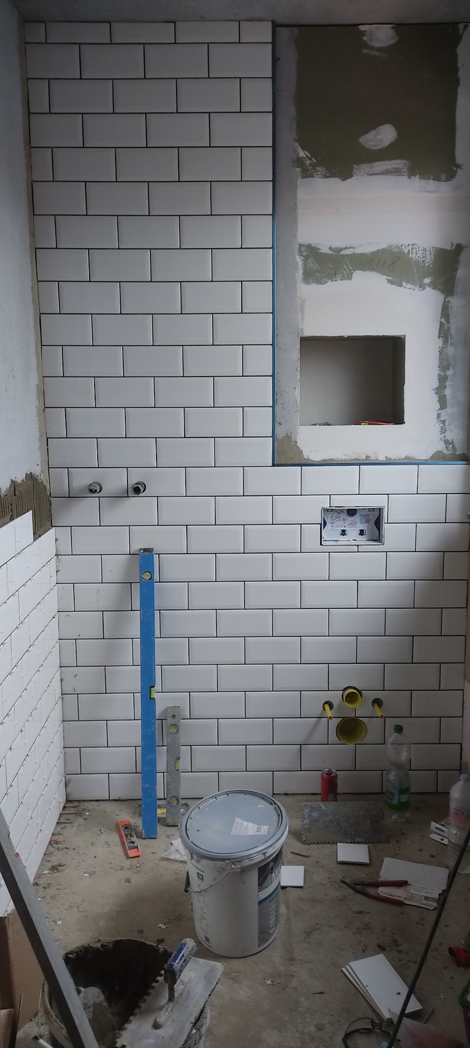 Układanie białych, prostokątnych płytek w łazience, widoczne narzędzia glazurnicze, poziomice, puszka instalacyjna, otwory na rury, częściowo wykończona ściana z wnęką.