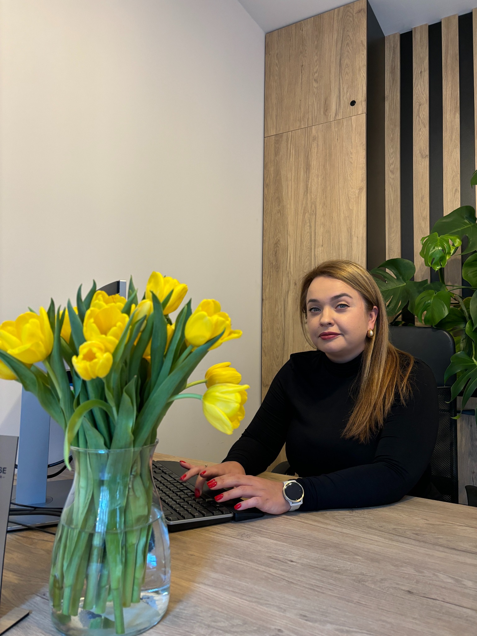 Kobieta w czarnej bluzce siedzi za biurkiem z laptopem, obok wazon z żółtymi tulipanami, w tle drewniane panele ścienne i zielona roślina doniczkowa.