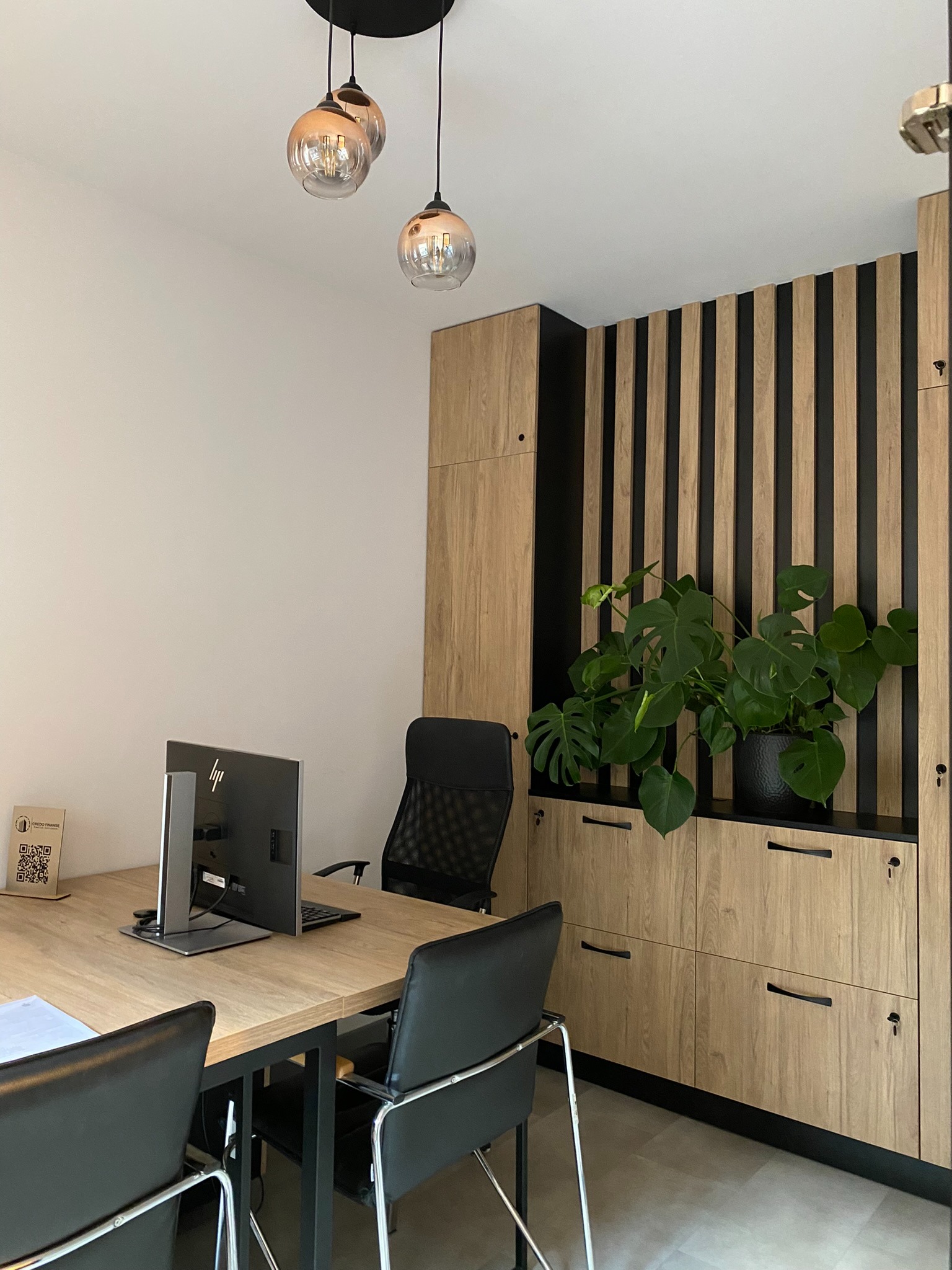 Nowoczesne biuro doradcy: biurko z komputerem, czarne krzesła, drewniane szafki z czarnymi akcentami, roślina doniczkowa, wiszące lampy z przezroczystymi kloszami.