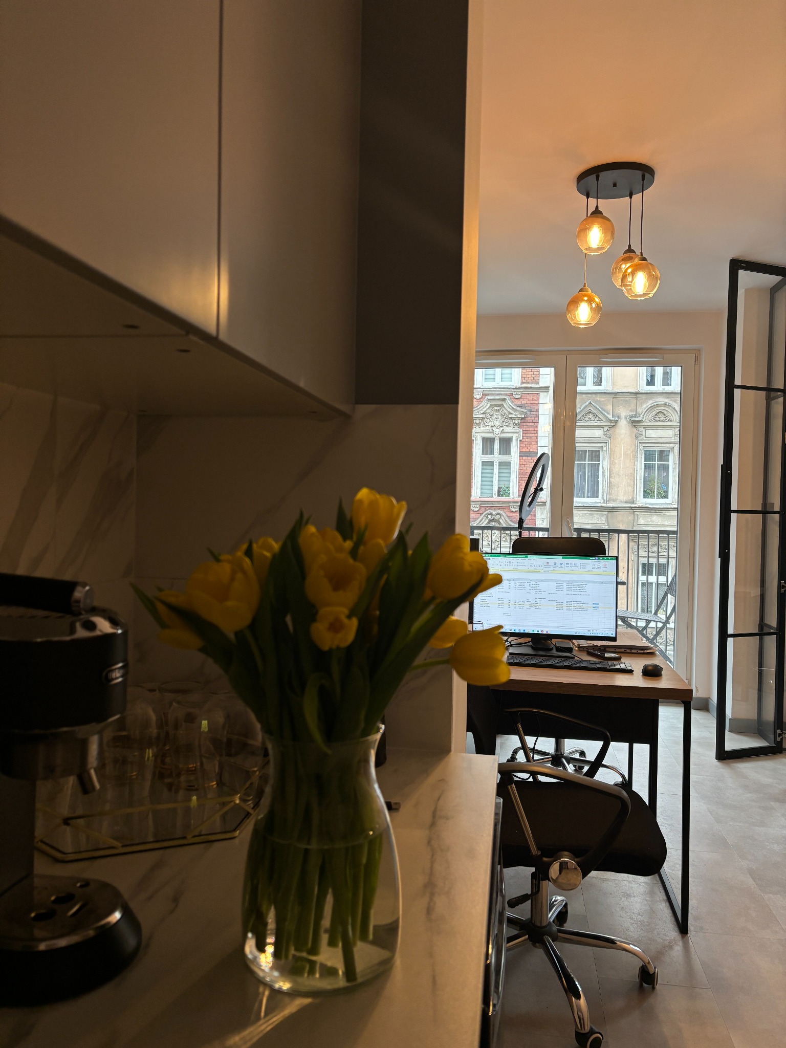 Nowoczesne biuro domowe z widokiem na kamienicę, żółte tulipany w wazonie na pierwszym planie, ekspres do kawy obok.