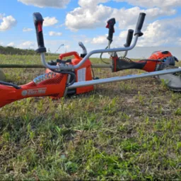 Dwie pomarańczowe podkaszarki spalinowe marki Oleo-Mac i Husqvarna leżą na trawie przed białym budynkiem magazynowym w słoneczny dzień.
