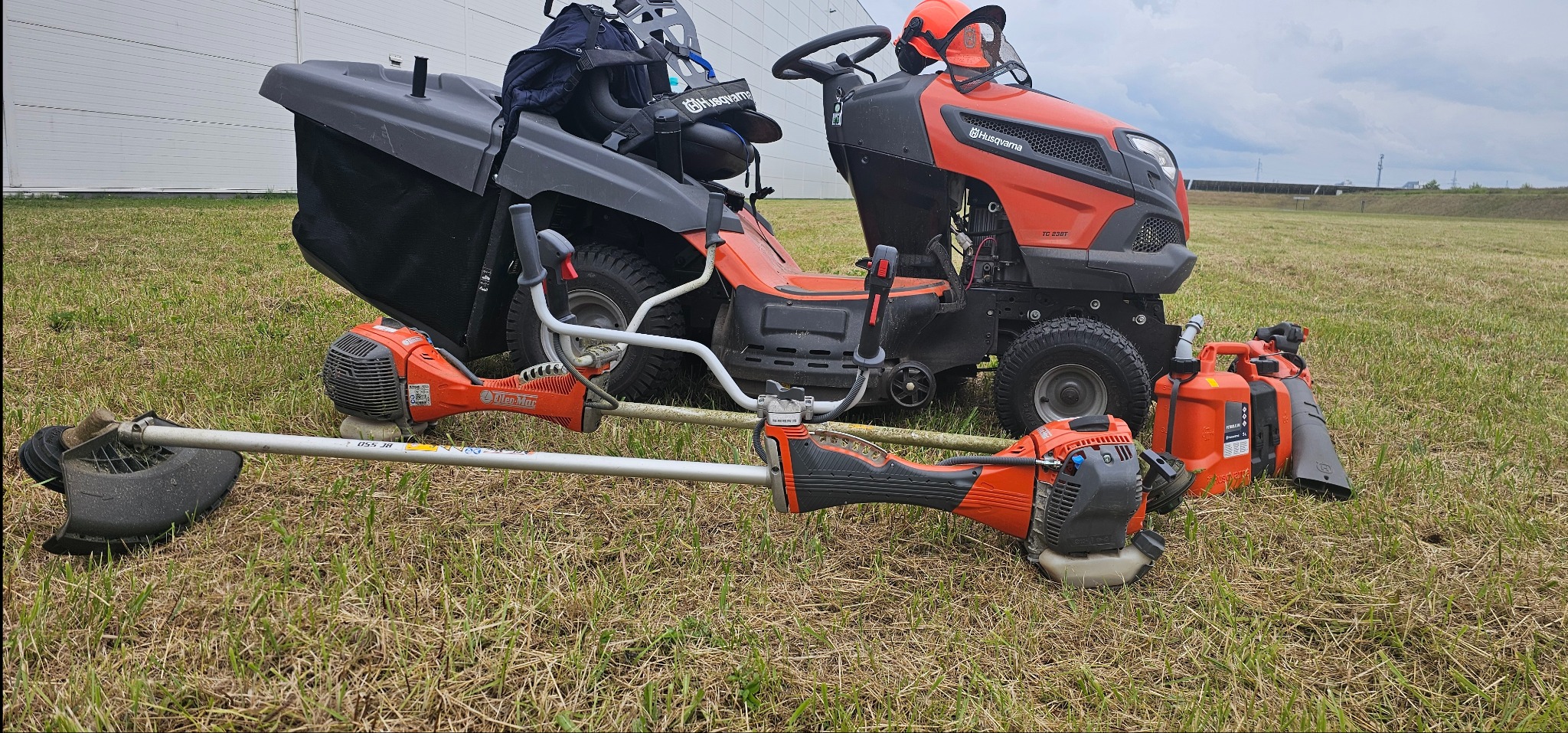 Pomarańczowy traktorek do koszenia trawy Husqvarna TC 238T i dwie podkaszarki spalinowe Husqvarna oraz kanister na tle zielonej trawy i hali.