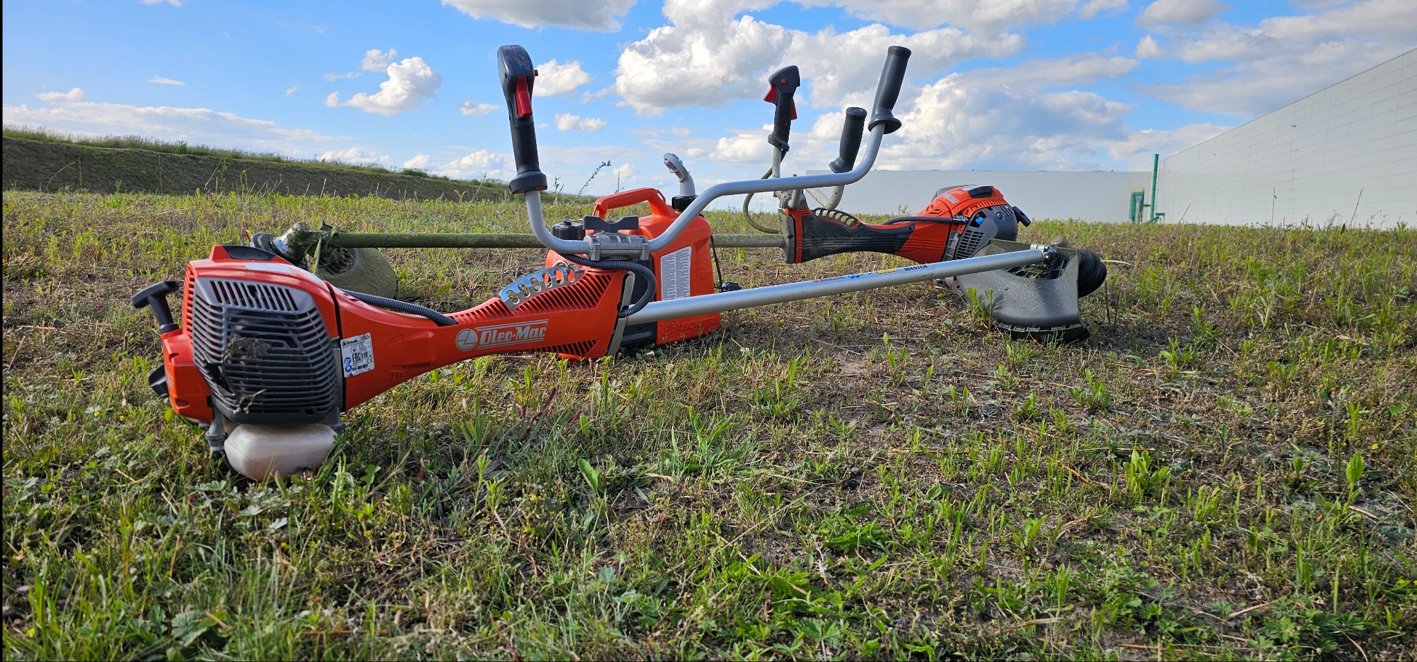 Dwie pomarańczowe podkaszarki spalinowe marki Oleo-Mac i Husqvarna leżą na trawie przed białym budynkiem magazynowym w słoneczny dzień.