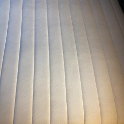 Pranie Tapicerki, konserwacja skór czyszczenie parowe, ozonowanie - Jasny materac z wyraźnymi, pionowymi przeszyciami, ukazujący czystą tapicerkę po profesjonalnym praniu. Widoczna faktura materiału i staranne wykończenie.