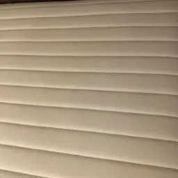 Jasny, prążkowany materac z delikatnymi wgłębieniami, ukazujący czystą powierzchnię i regularny wzór. Widoczny fragment ciemnego zagłówka.