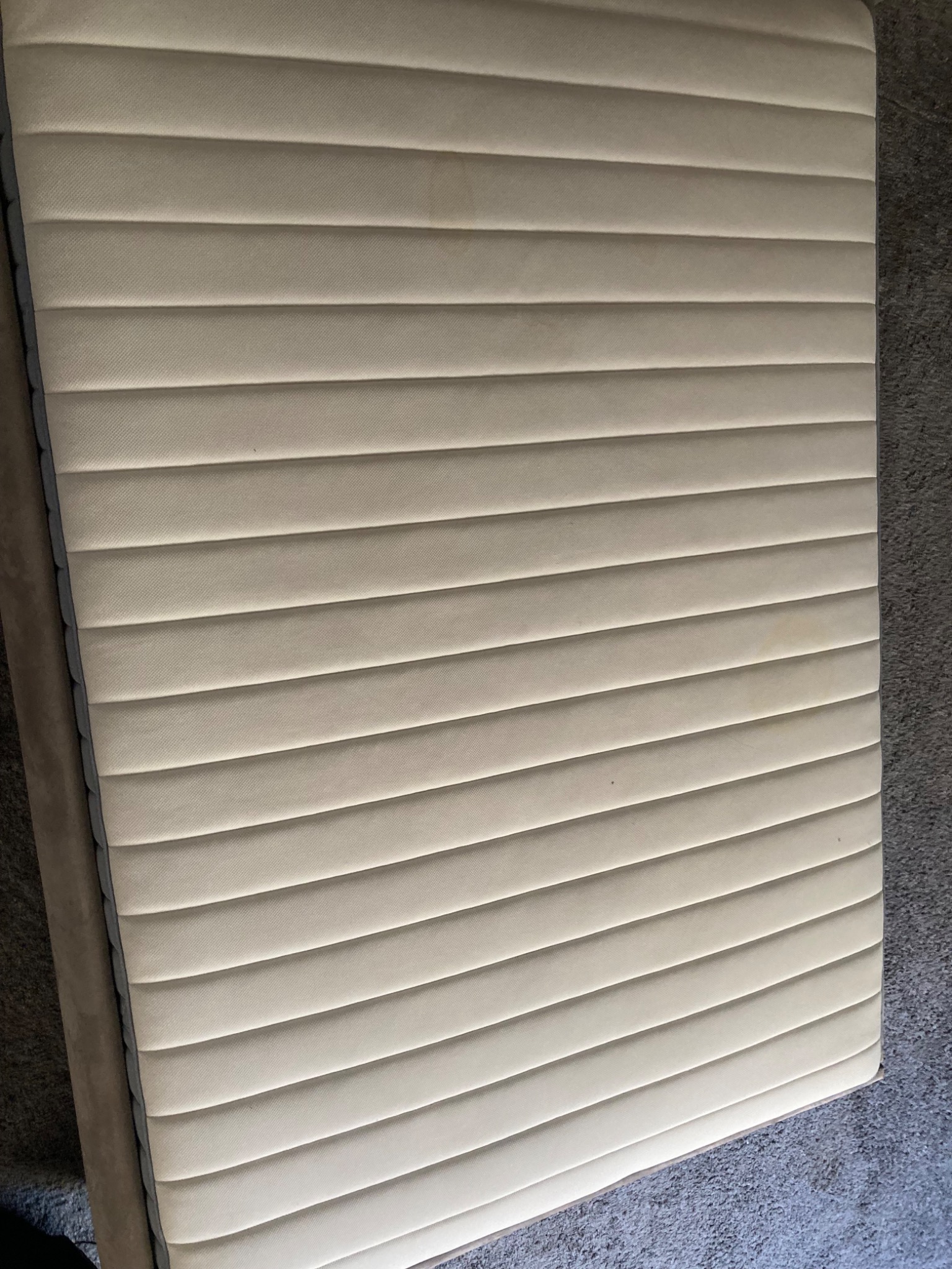 Beżowy materac z wyraźnymi, poziomymi przeszyciami leży na szarym dywanie. Widoczne plamy na powierzchni materaca.