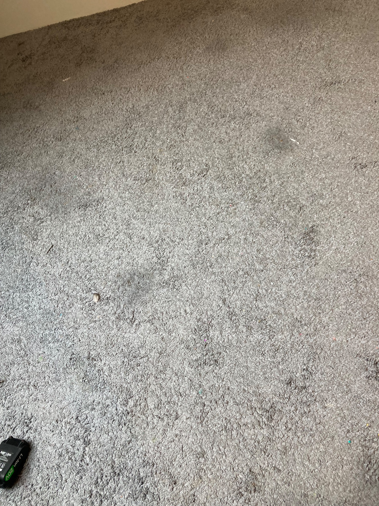 Szary dywan z widocznymi plamami i zanieczyszczeniami, bateria urządzenia czyszczącego w rogu. Dywan wymaga odświeżenia.