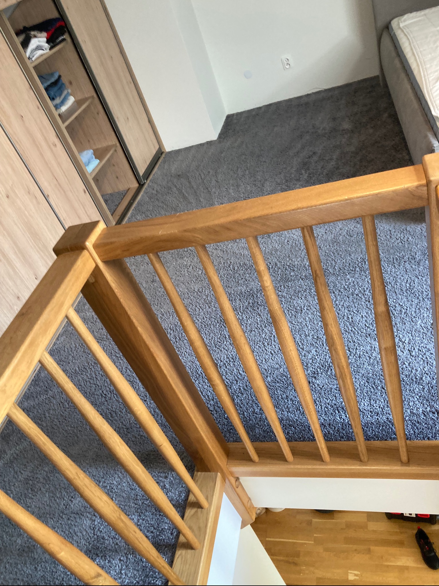Szary dywan na schodach, widok z góry przez drewnianą balustradę. W tle szafa z ubraniami i fragment łóżka. Tekstura dywanu wyraźnie widoczna.