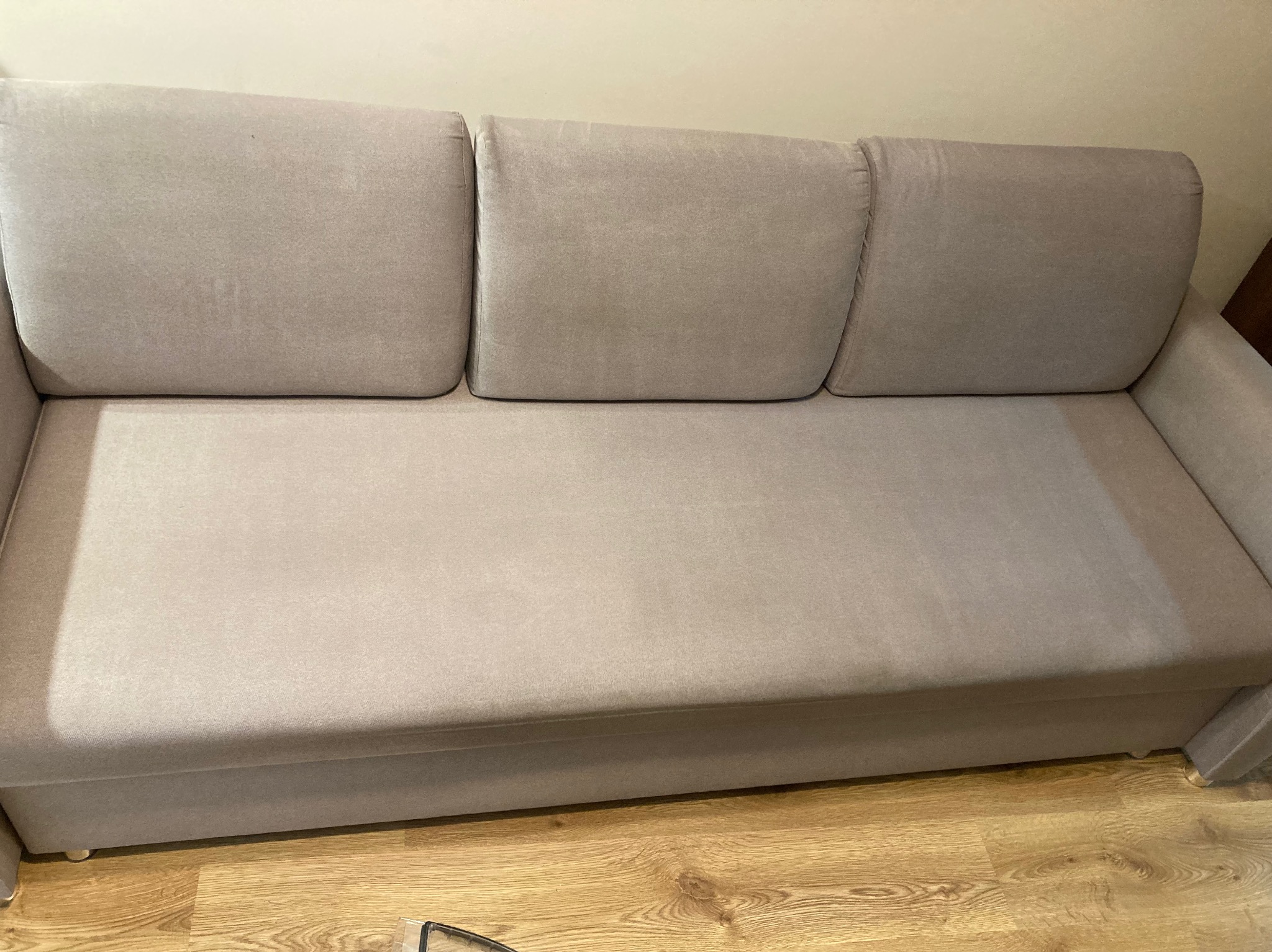 Jasnoszara sofa z trzema poduchami na oparciu, widoczne plamy na tapicerce, drewniana podłoga. Ujęcie z góry, naturalne światło.