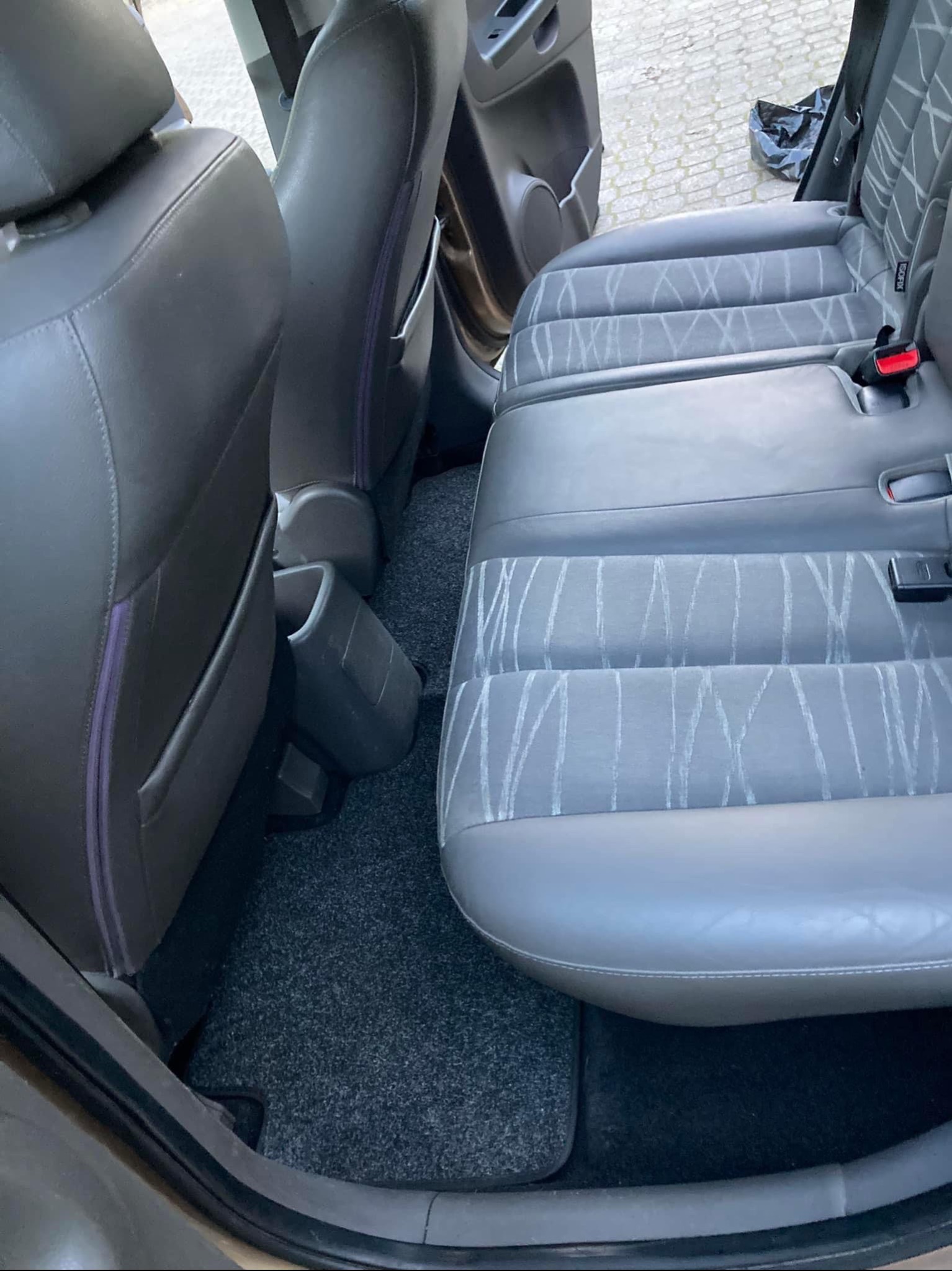 Szare, tylne siedzenie samochodu z wzorem w jasne linie, widoczne mocowania ISOFIX i dywanik podłogowy. Ujęcie z wnętrza pojazdu, z otwartymi drzwiami.