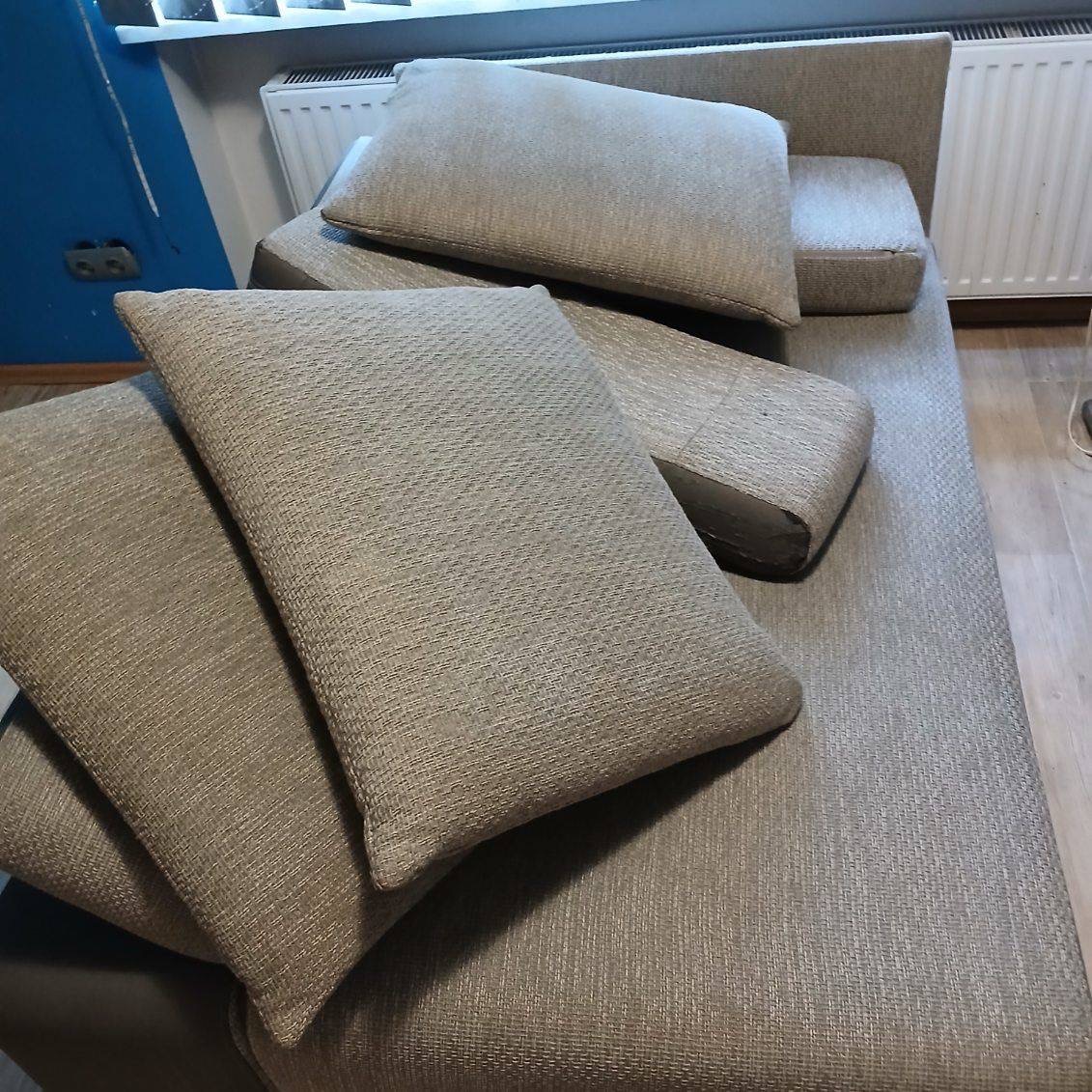 Jasnoszara kanapa z poduszkami o wyraźnej fakturze materiału, ustawiona w pomieszczeniu z niebieską ścianą i grzejnikiem. Widoczne detale tapicerki i układ poduszek.