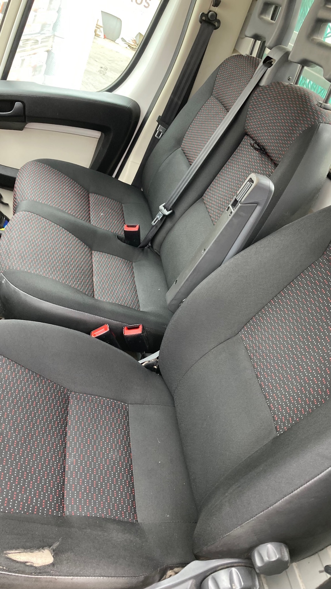 Wnętrze busa z tapicerowanymi siedzeniami w odcieniach szarości z czerwonym wzorem. Widoczne pasy bezpieczeństwa i podłokietnik. Ujęcie z perspektywy pasażera.