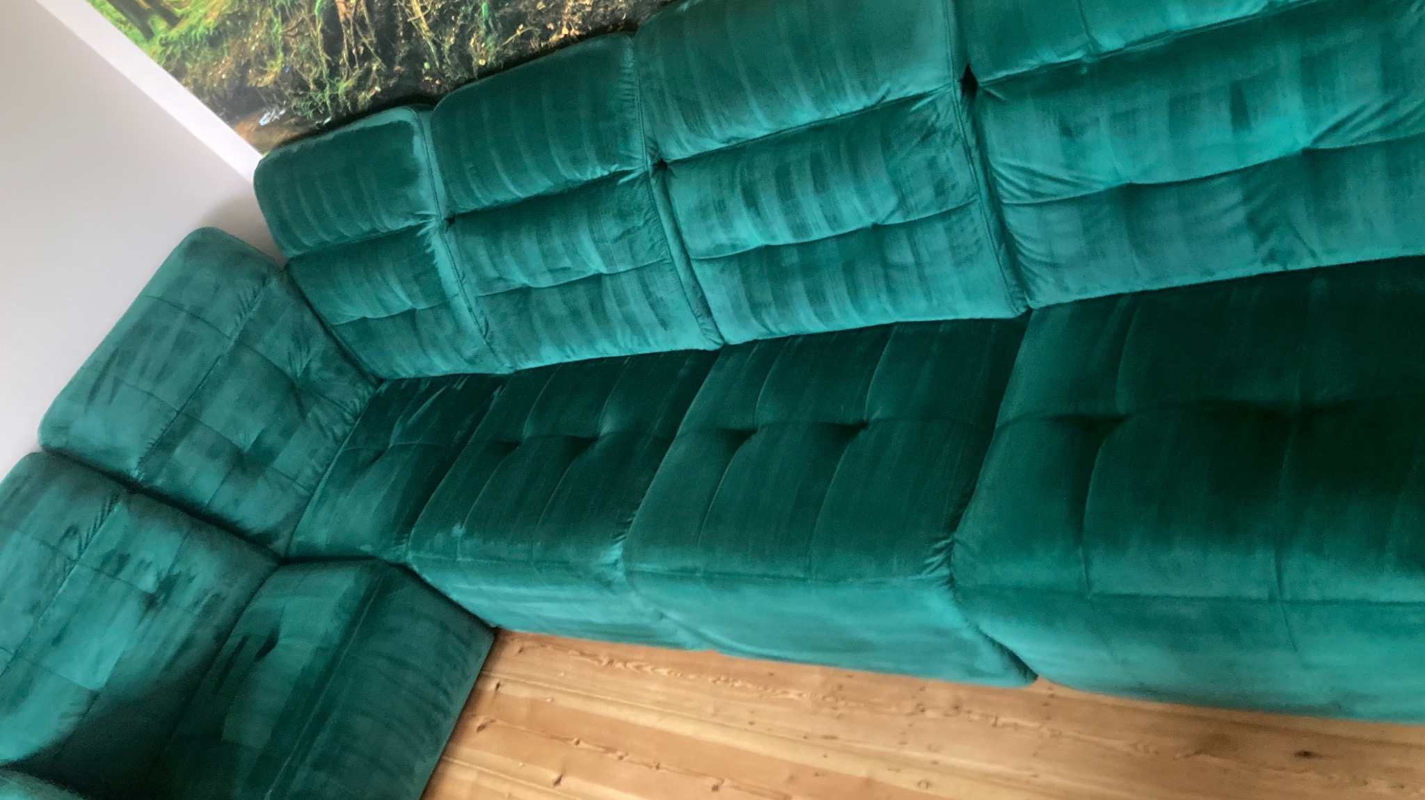 Szmaragdowa, modułowa sofa z aksamitną tapicerką, widoczna z góry, na drewnianej podłodze. W tle fragment dekoracji ściennej z motywem leśnym.