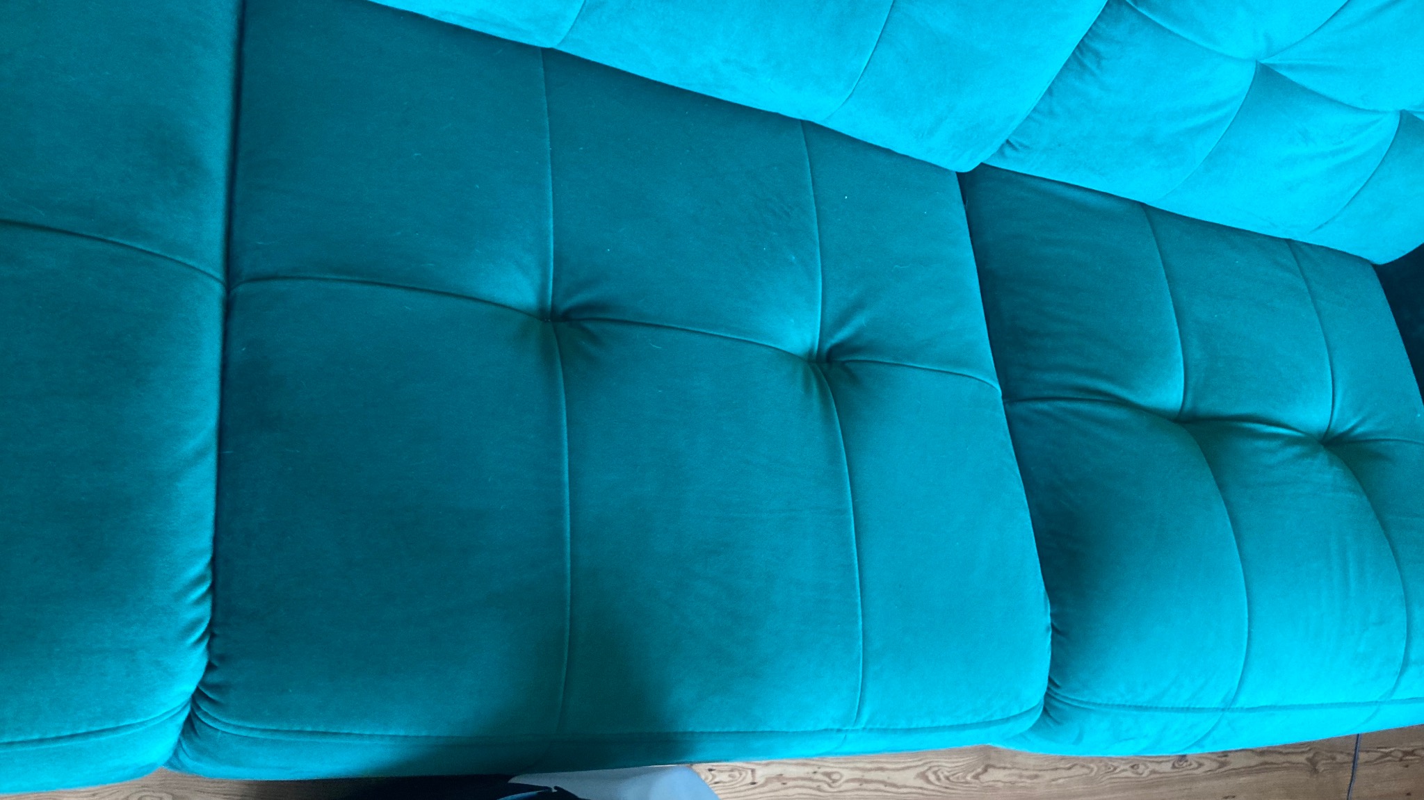 Szmaragdowa, aksamitna sofa z głębokimi pikowaniami, widziana z góry, ukazuje miękkość i komfort mebla. Widoczny fragment drewnianej podłogi dodaje ciepła.