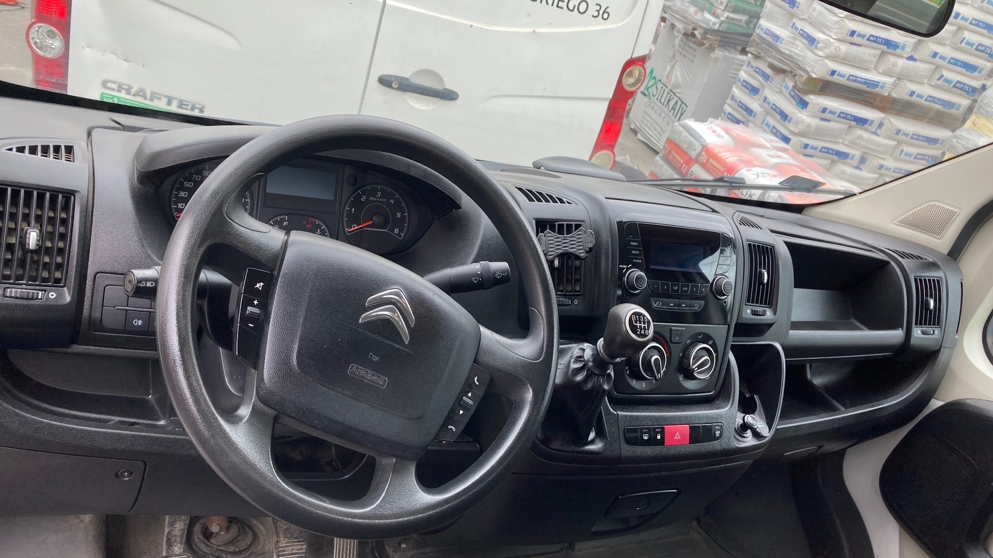 Wnętrze kabiny busa marki Citroen z widoczną deską rozdzielczą, kierownicą z logo, dźwignią zmiany biegów i stertą worków z zaprawą budowlaną w tle.