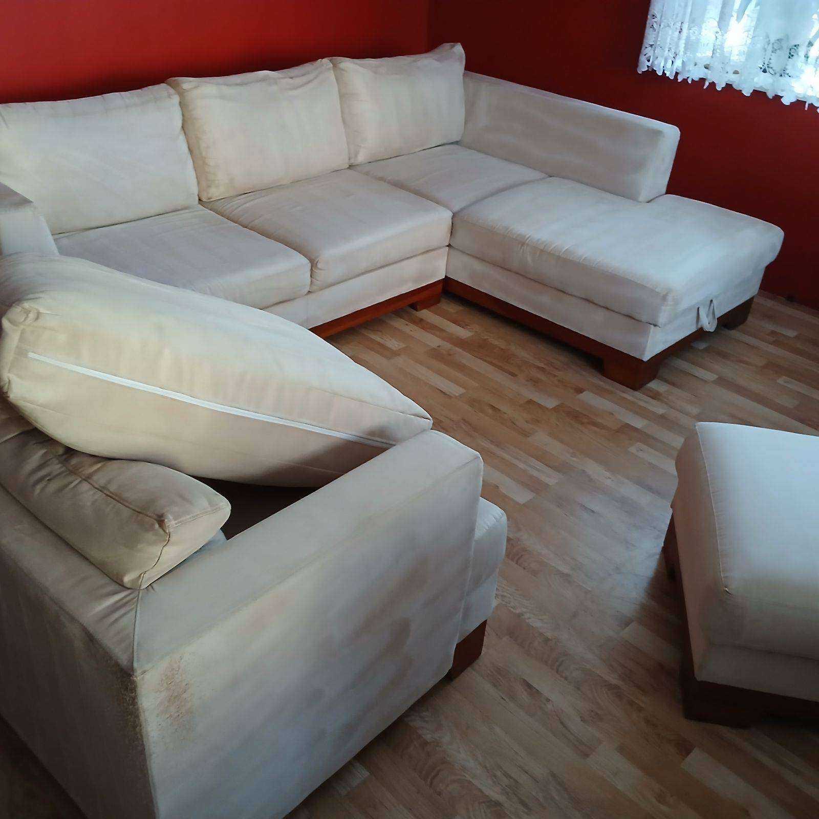 Beżowy narożnik z widocznymi plamami na tapicerce, ustawiony na drewnianej podłodze. Widoczny również puf. Czerwona ściana w tle.