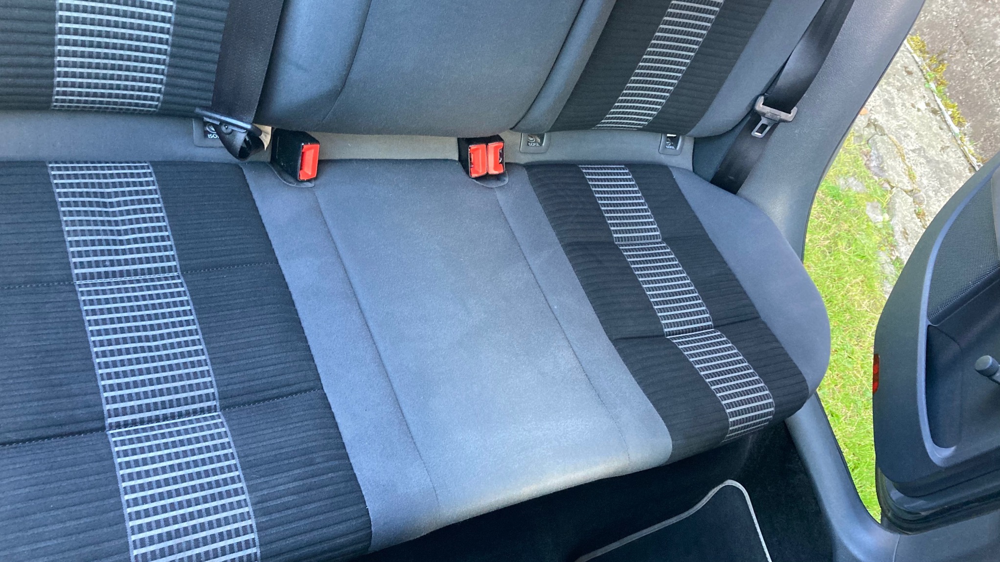 Szara, tekstylna tylna kanapa samochodowa z pasami bezpieczeństwa i mocowaniami ISOFIX, widoczna z bliska, z fragmentem otwartych drzwi i zielenią w tle.