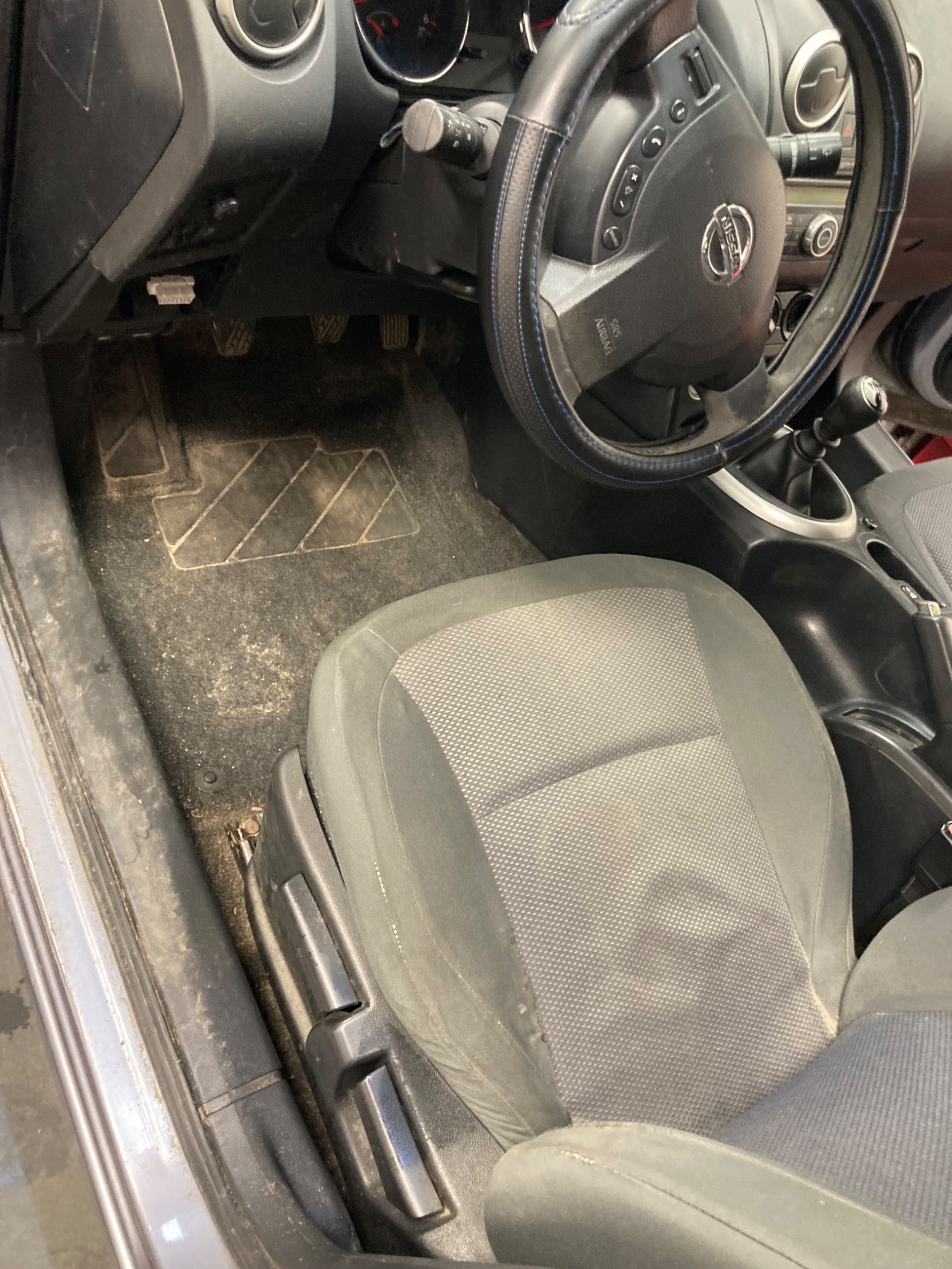 Brudne wnętrze samochodu Nissan: fotel kierowcy z plamami, dywaniki z piaskiem i kurzem, kierownica w pokrowcu. Wymaga gruntownego czyszczenia.