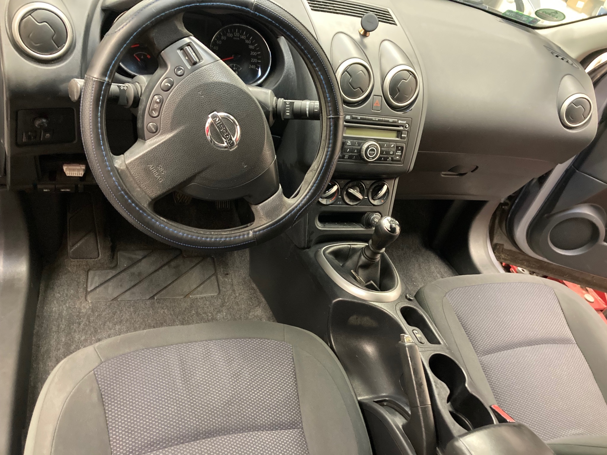 Wnętrze samochodu Nissan z widocznymi szarymi fotelami, kierownicą z niebieskimi przeszyciami i deską rozdzielczą. Widok z perspektywy pasażera.