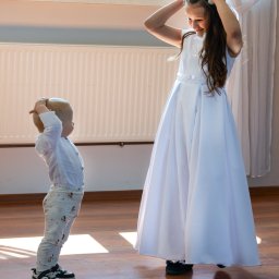 Photolita_studio - Dziewczynka w białej sukience i wianku na głowie, poprawia go, a mały chłopiec w koszuli i spodniach z nadrukiem, naśladuje ją, stojąc na drewnianej podłodze w jasnym wnętrzu.