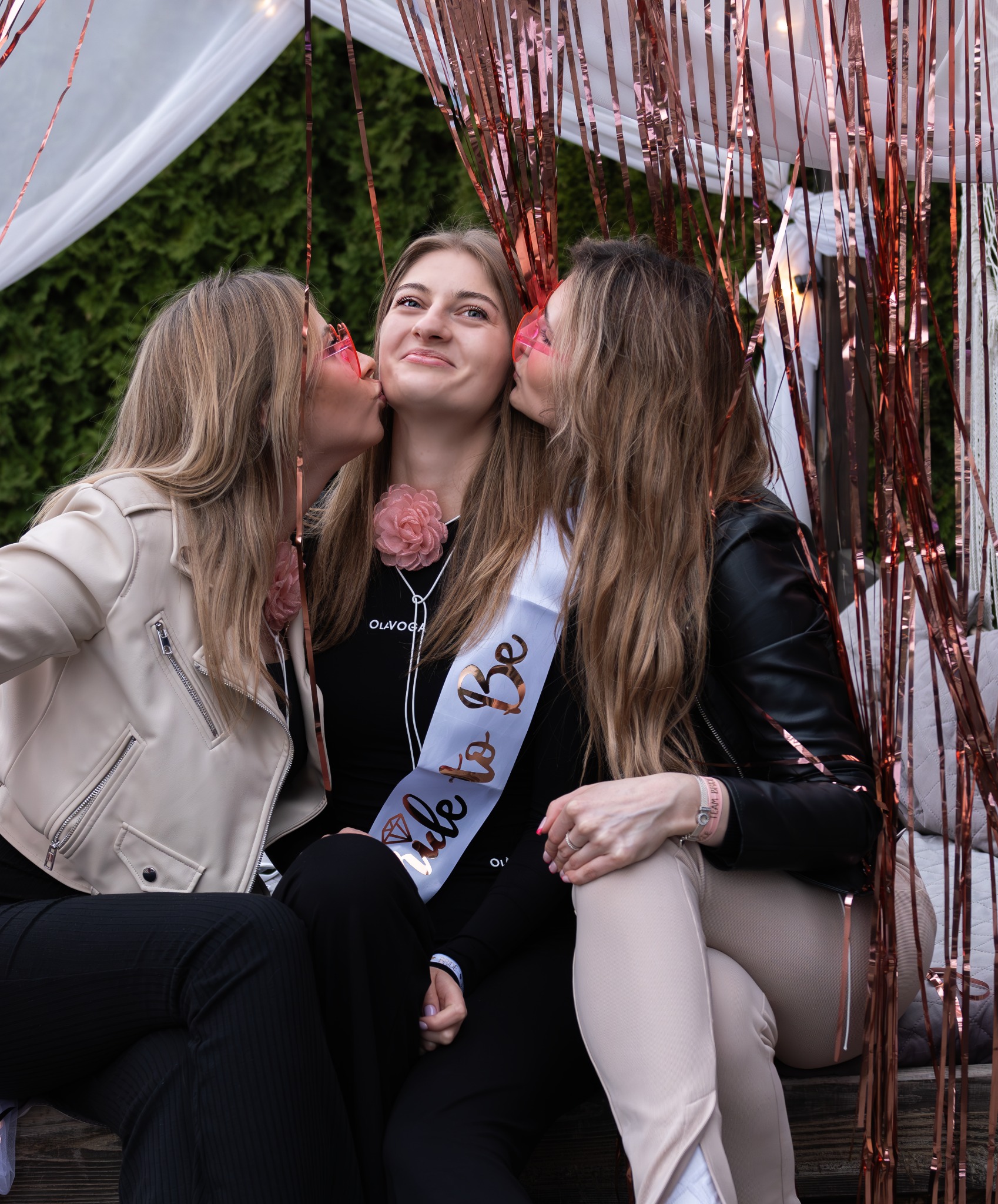 Trzy uśmiechnięte kobiety w plenerze, panna młoda w szarfie 'Bride to Be' z kwiatową ozdobą, całowana przez dwie przyjaciółki w różowych okularach.